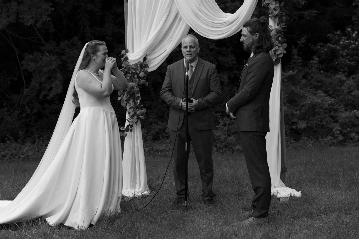 Black and white photo of an intimate outdoor wedding ceremony taking place under an elegant draped arch.