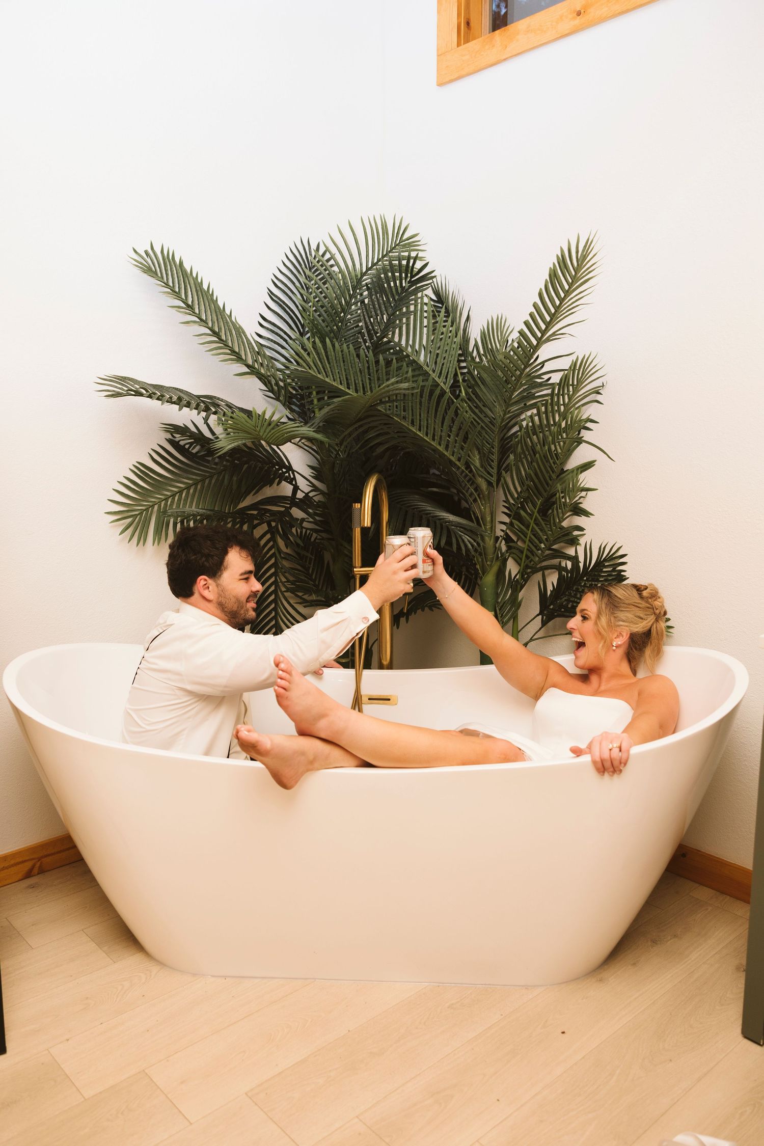 A couple shares a playful moment in a modern freestanding white bathtub with tropical plants in the background.
