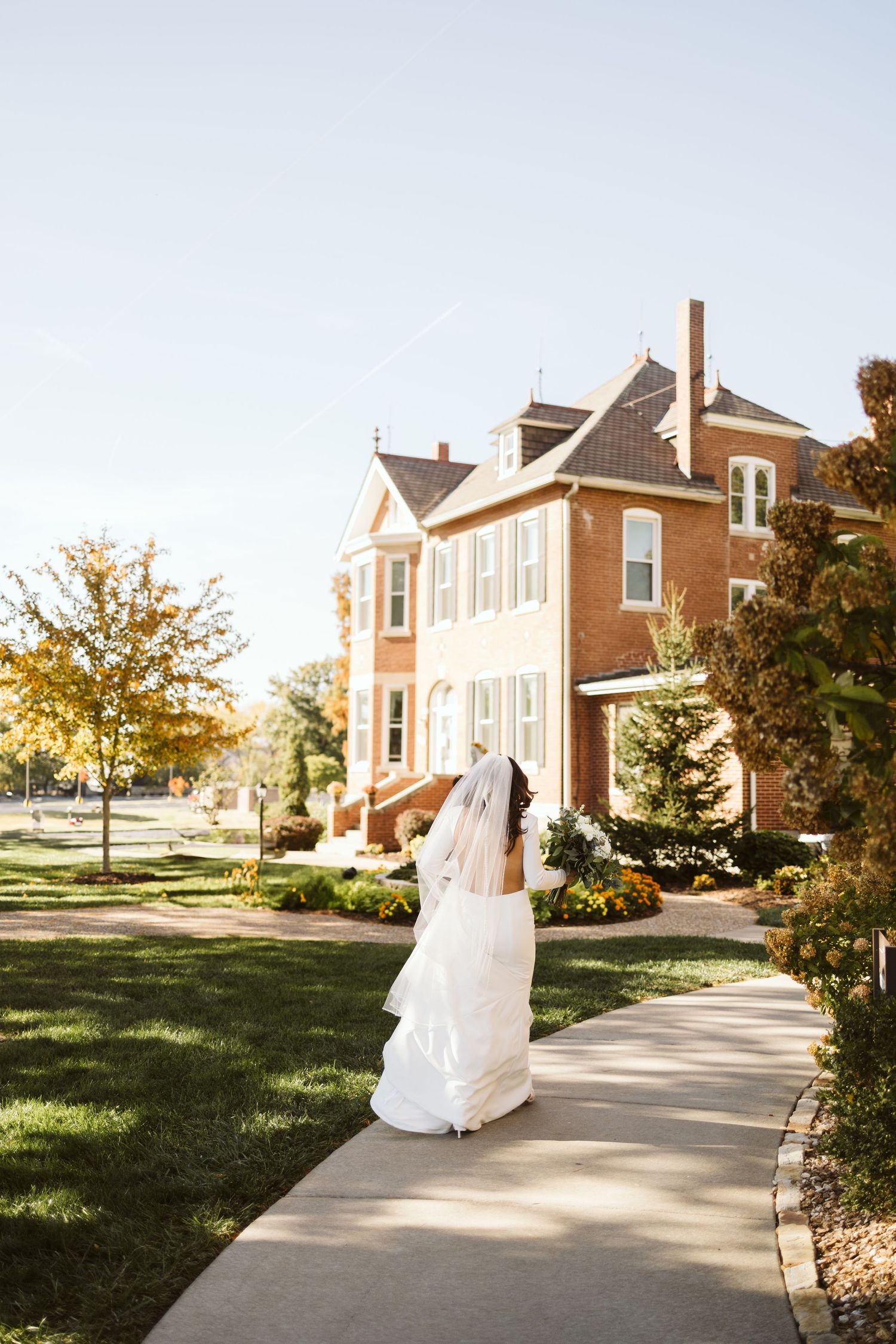 A bride in a flowing white dress walks along a path beside a historic brick building.