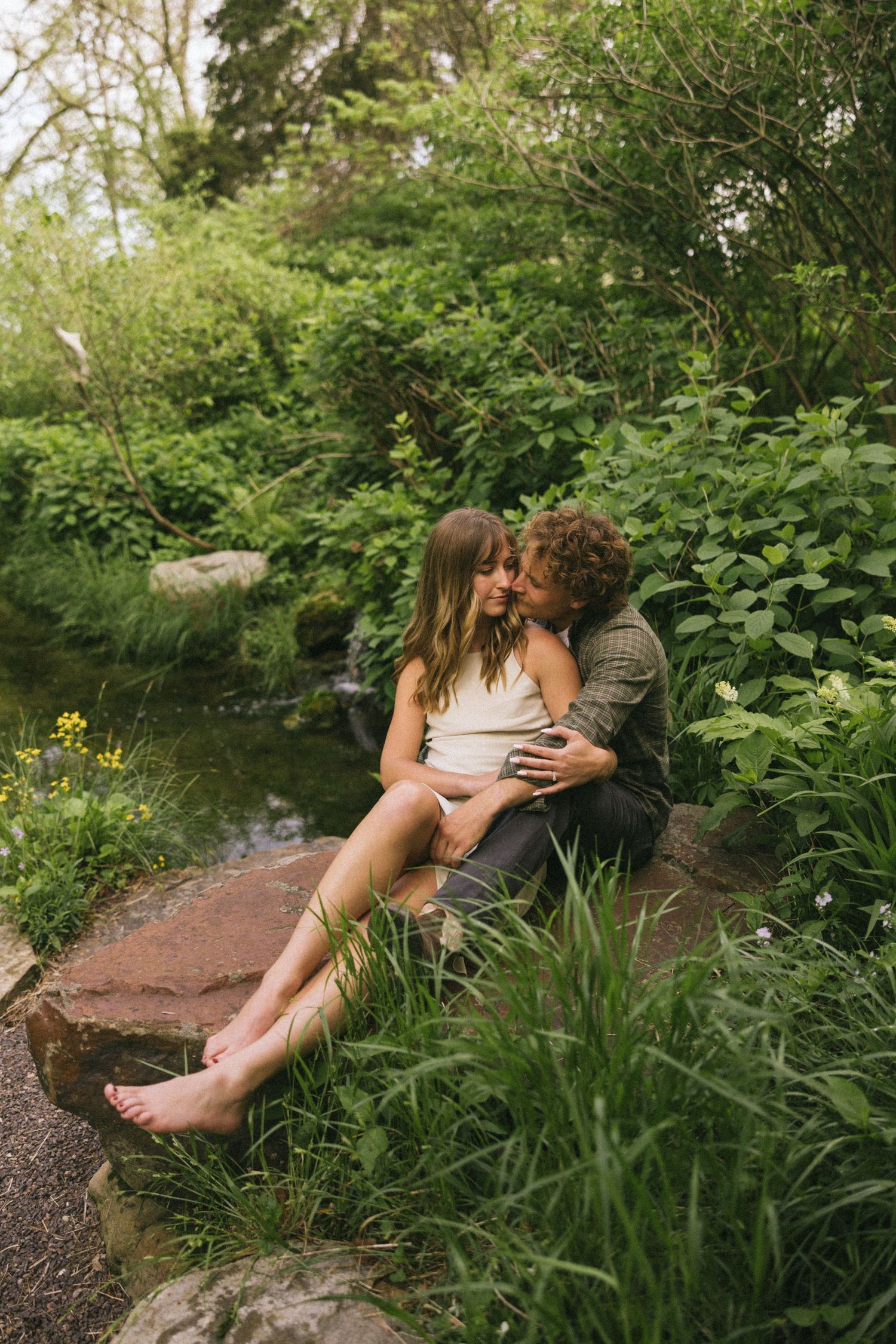 Two people share romantic moments on a stone ledge surrounded by garden plants and flowers.