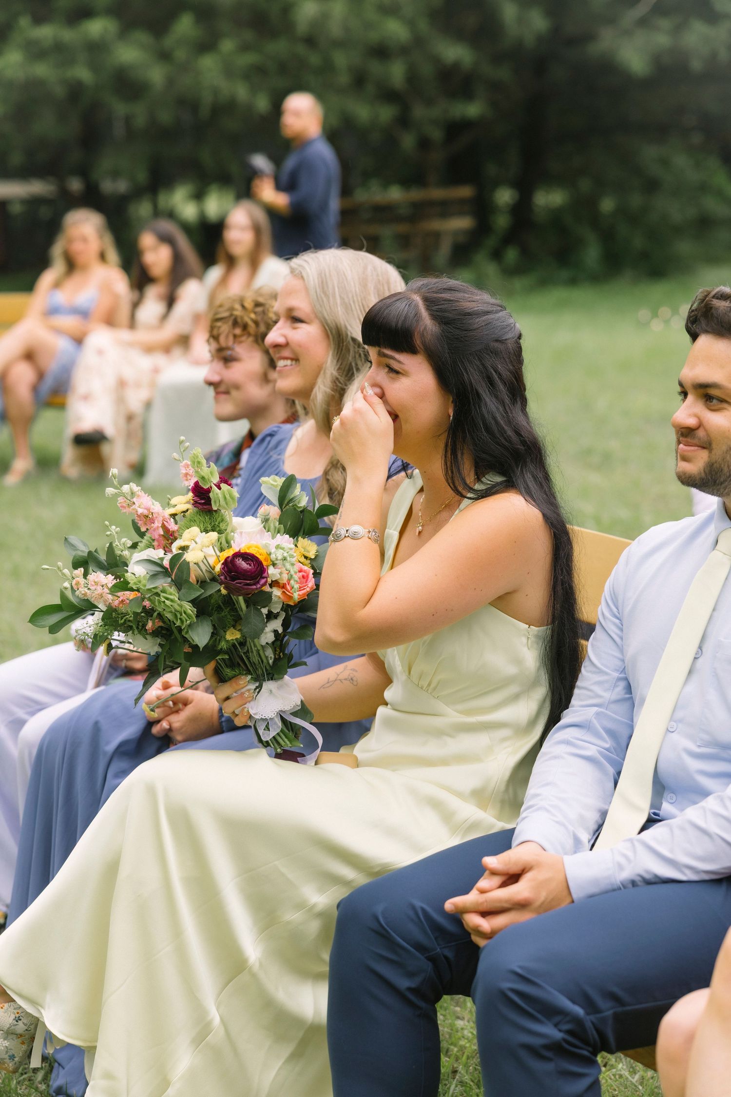 Wedding guests seated outdoors holding bouquets and sharing laughs during a ceremony.