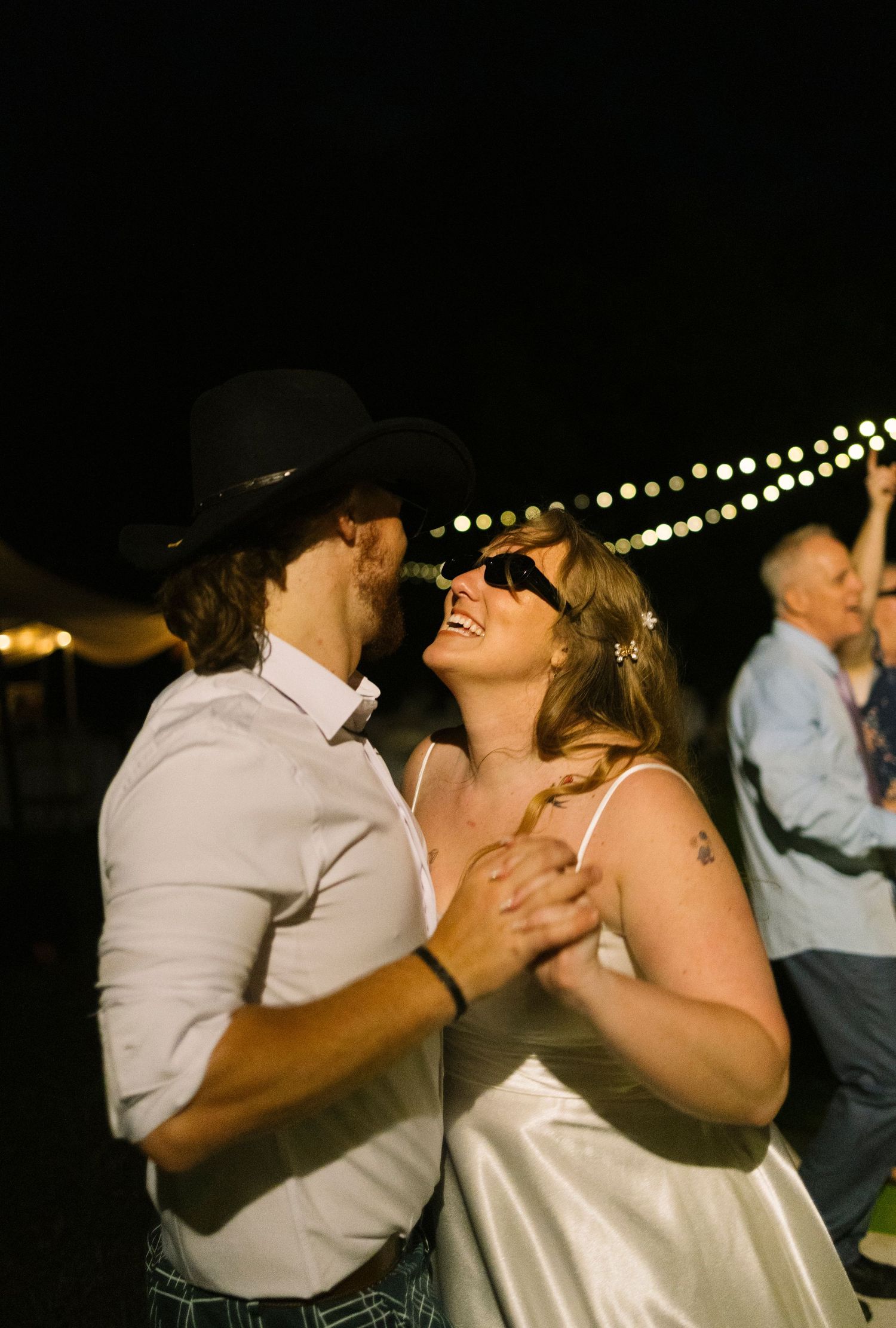 A couple shares a joyful dance moment under twinkling string lights at a nighttime outdoor celebration.