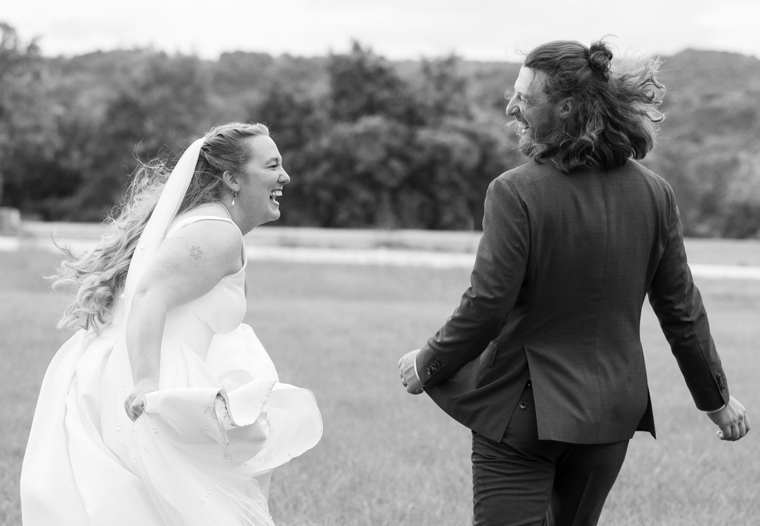 A joyful black and white photo showing two people running together in a field, with one wearing a wedding dress.