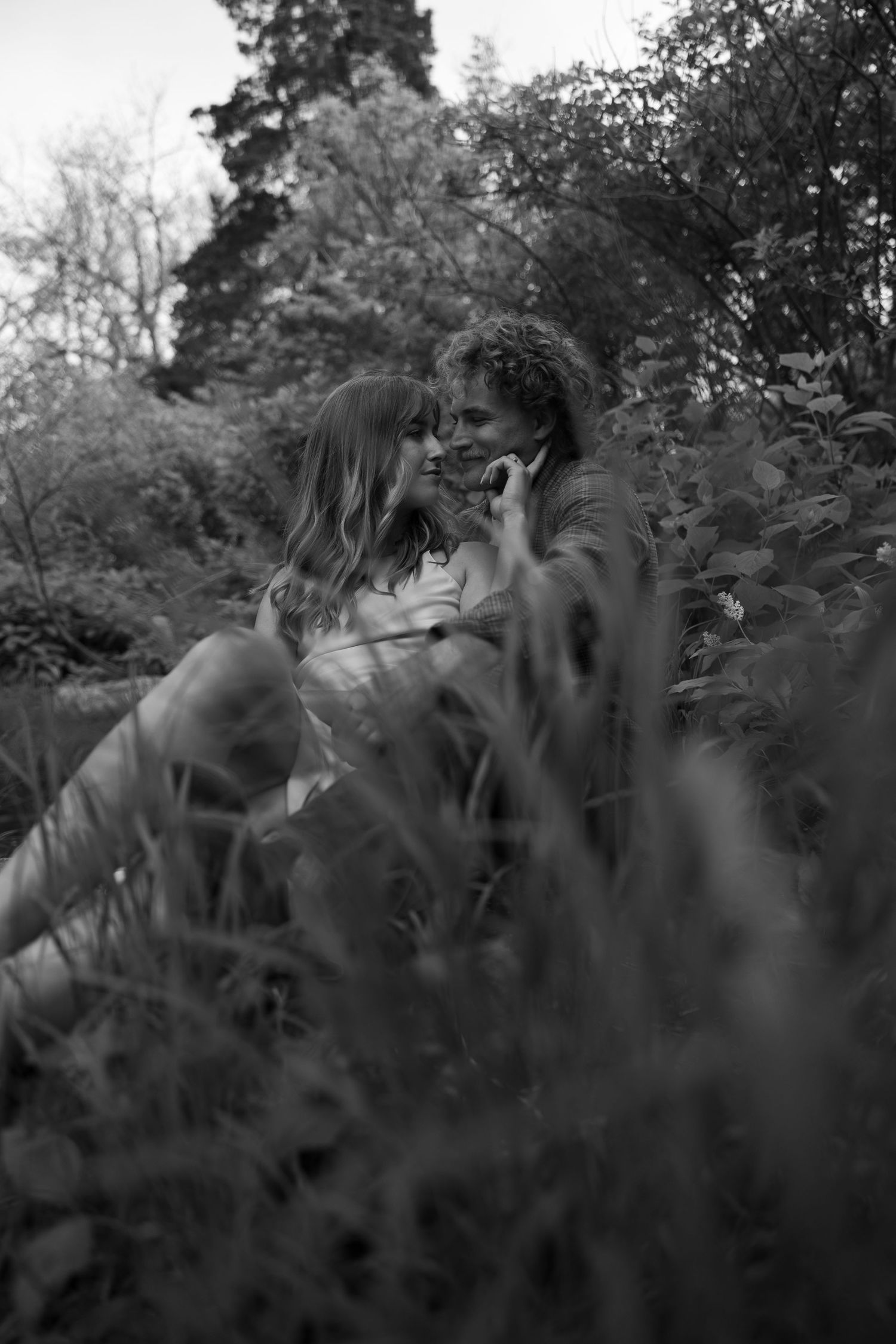 A black and white photo of a couple sharing an intimate moment among tall grass and foliage.