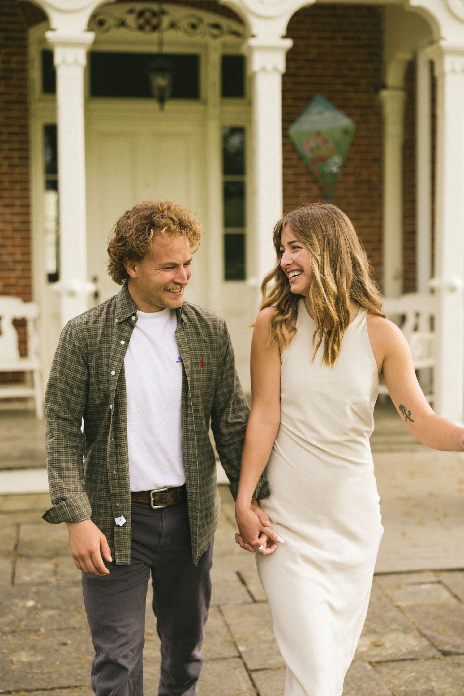 A couple holding hands and laughing while walking in front of a Victorian porch with white columns.