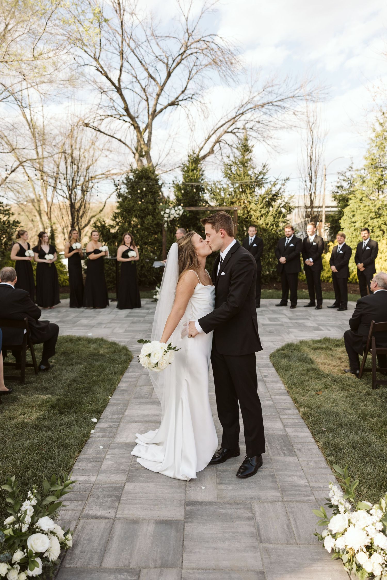 Elegant outdoor wedding ceremony at sunset with formal wedding party lined up along stone pathway adorned with flowers.