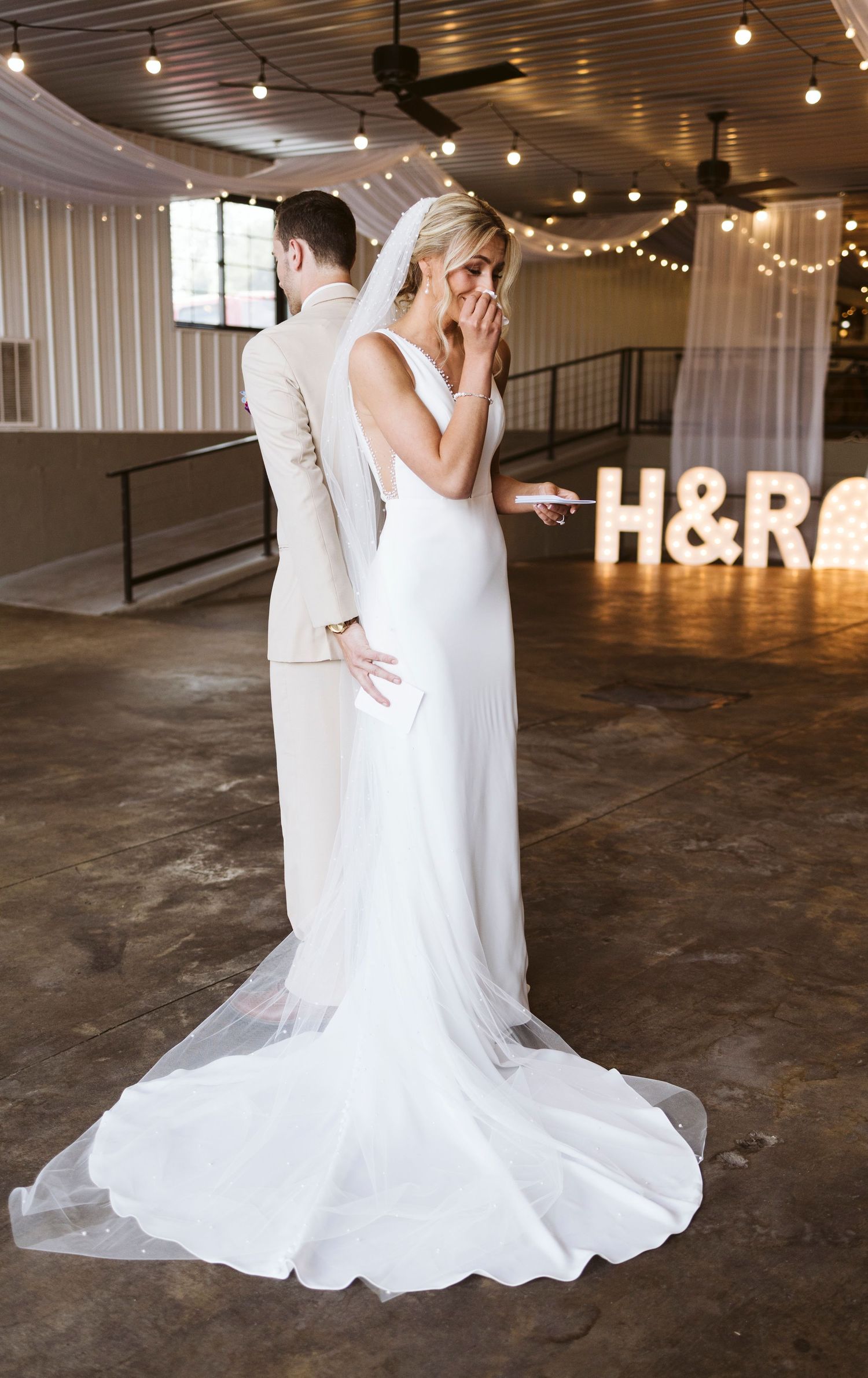 A bride in an elegant white gown poses in an industrial venue with string lights and illuminated initials.