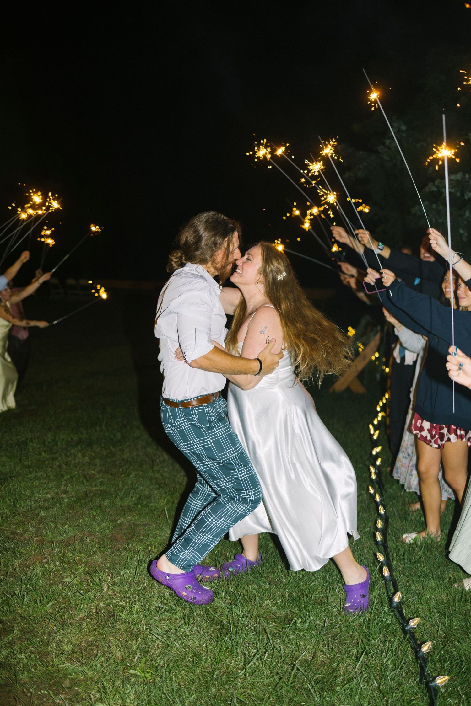 Group celebrating with sparklers at night on grassy field under dark sky with glowing lights.
