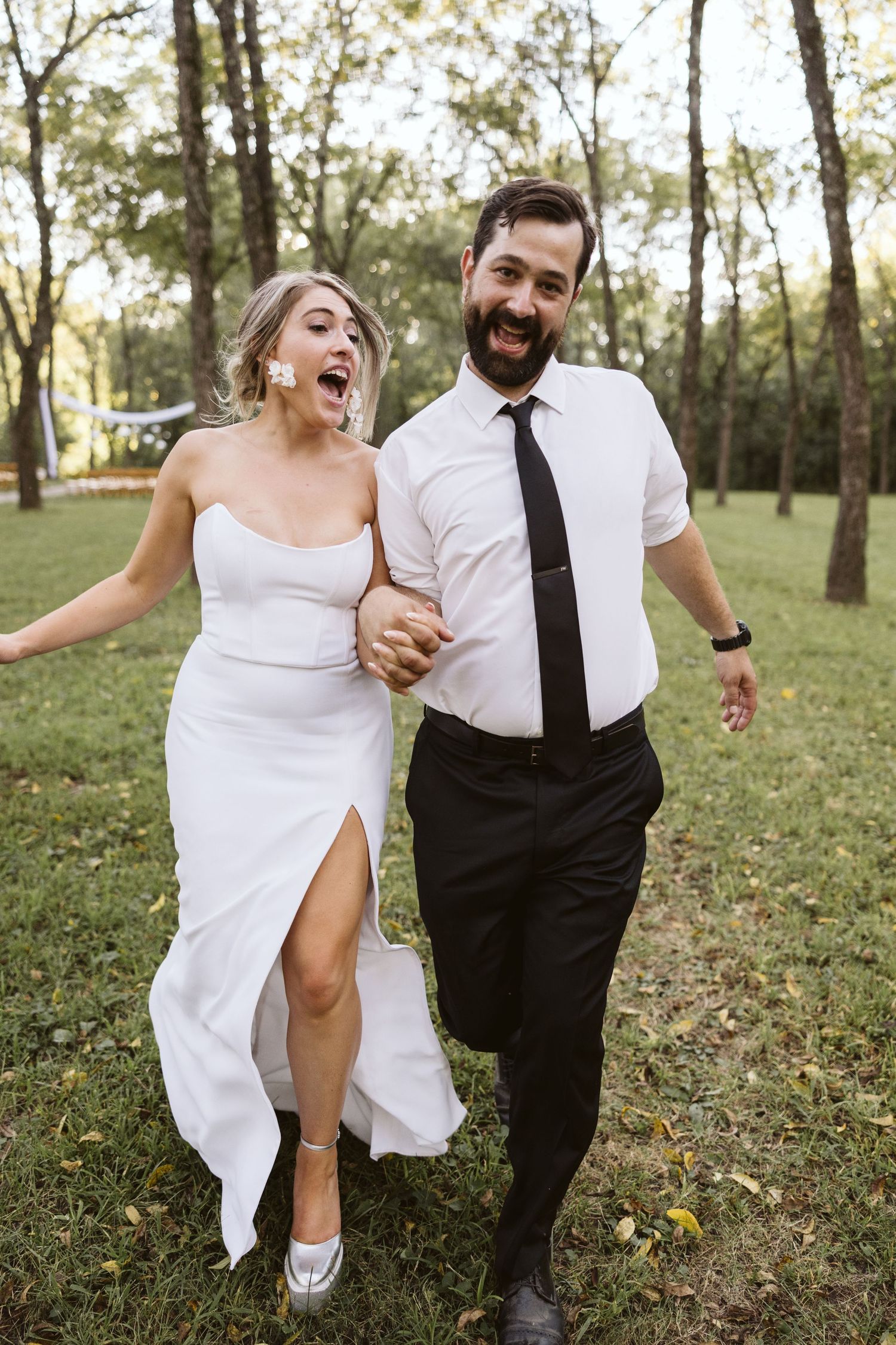 Wedding couple laughing and running playfully through a sunny wooded area.