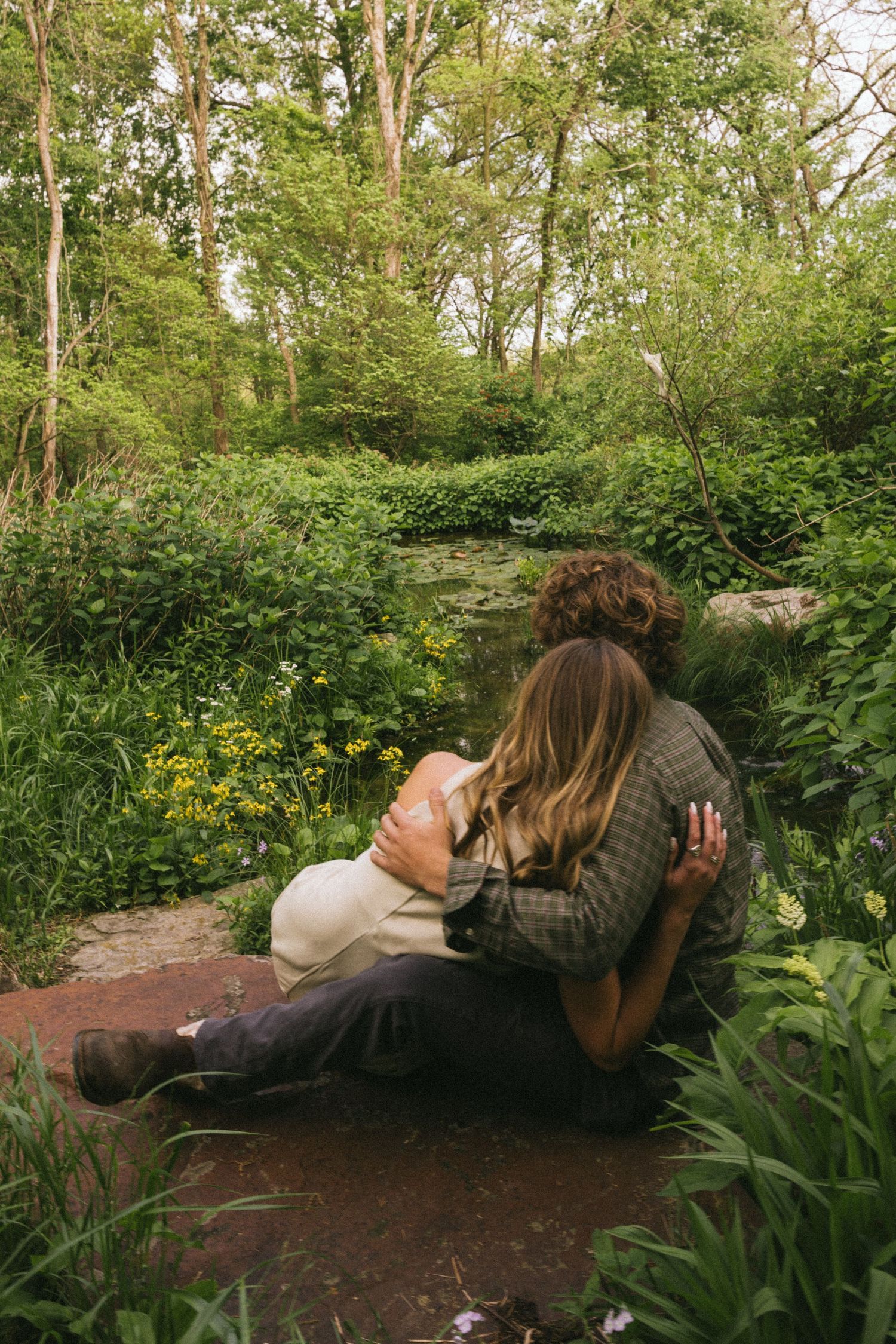 A couple sits together on a rock surrounded by lush green forest vegetation and yellow wildflowers.