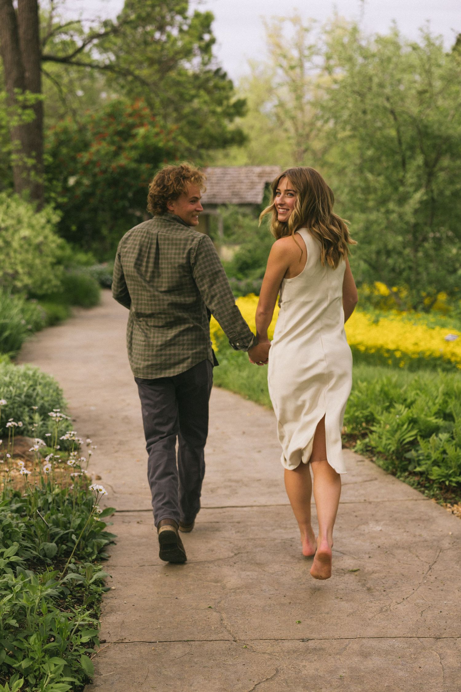 A romantic couple walking hand in hand on a garden path surrounded by spring flowers and trees.