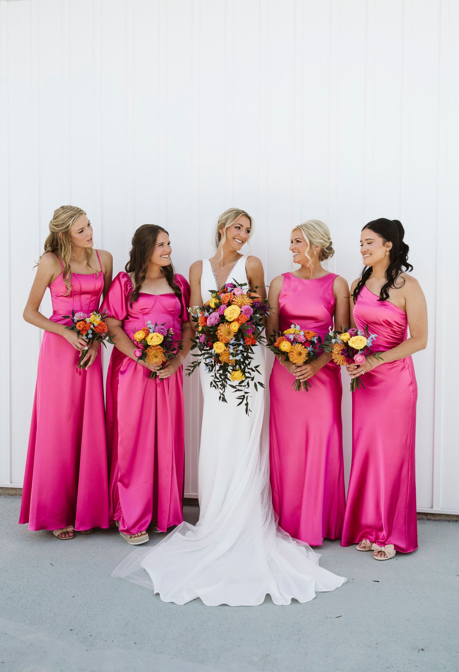 Bridesmaids in vibrant pink dresses holding colorful bouquets pose together against a white wall.