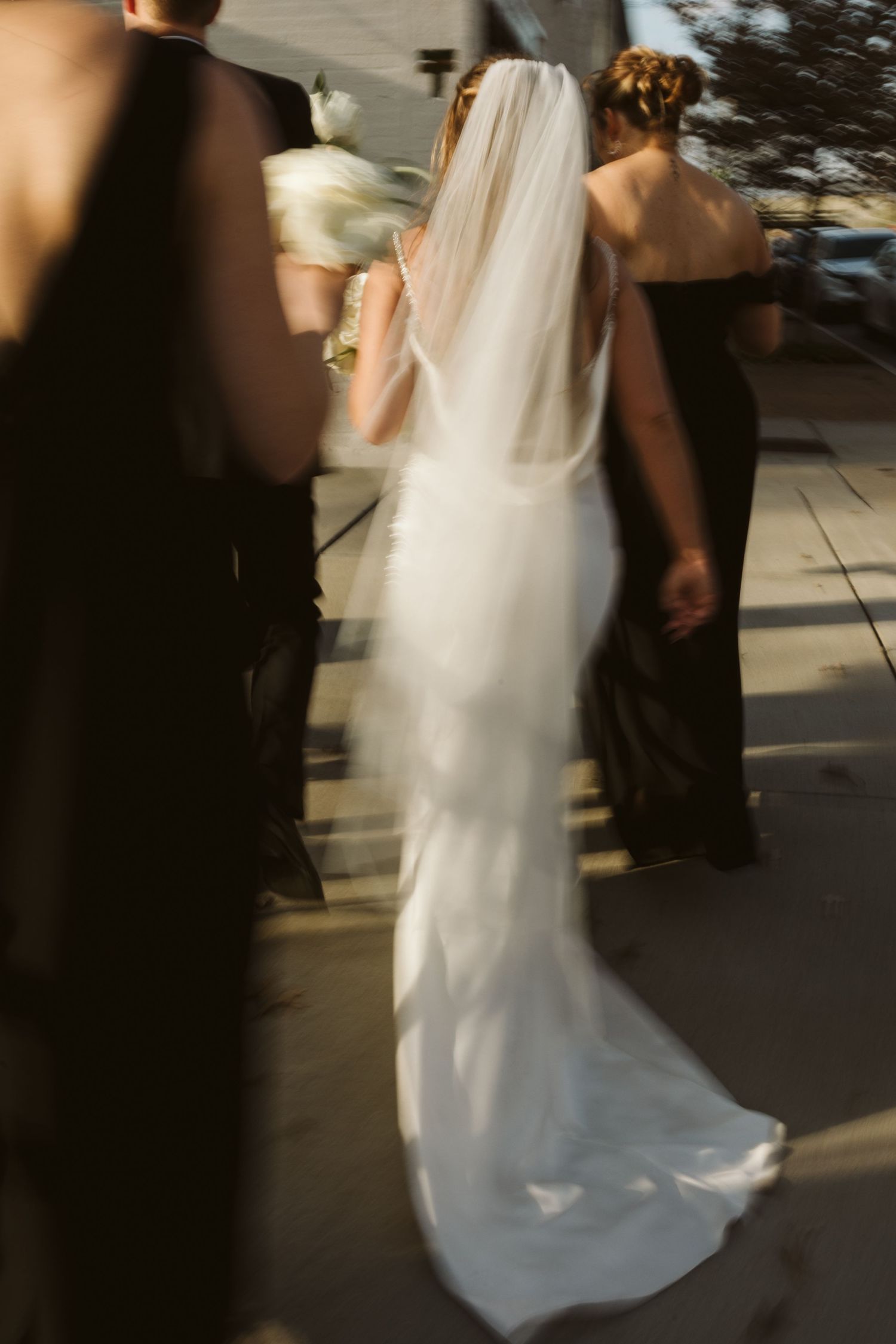 A bride in a flowing white wedding dress with a long veil walks along a pathway during a wedding ceremony.