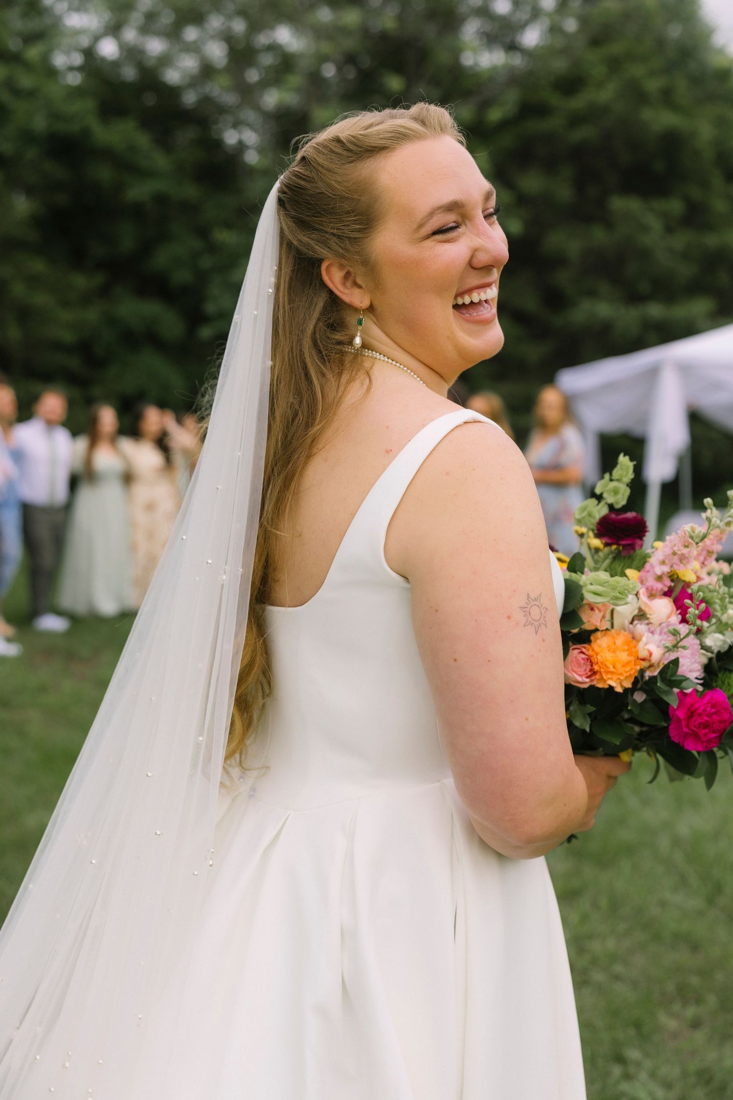 A candid portrait of a bride laughing while holding a colorful bouquet in an outdoor garden setting.