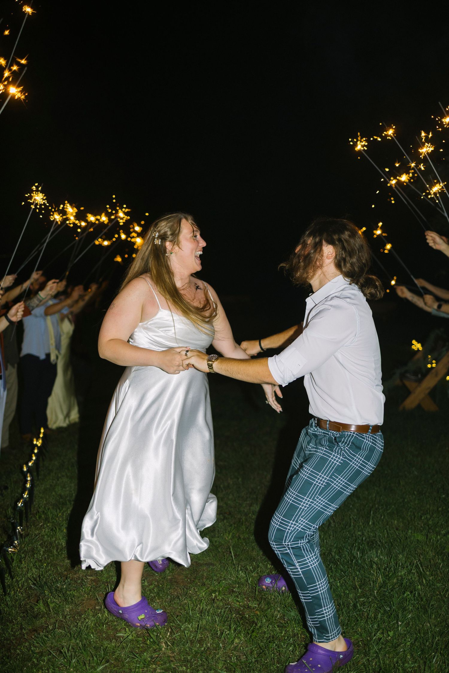 Sequence of four nighttime photos showing two people playfully dancing on grass with illuminated branches in background.