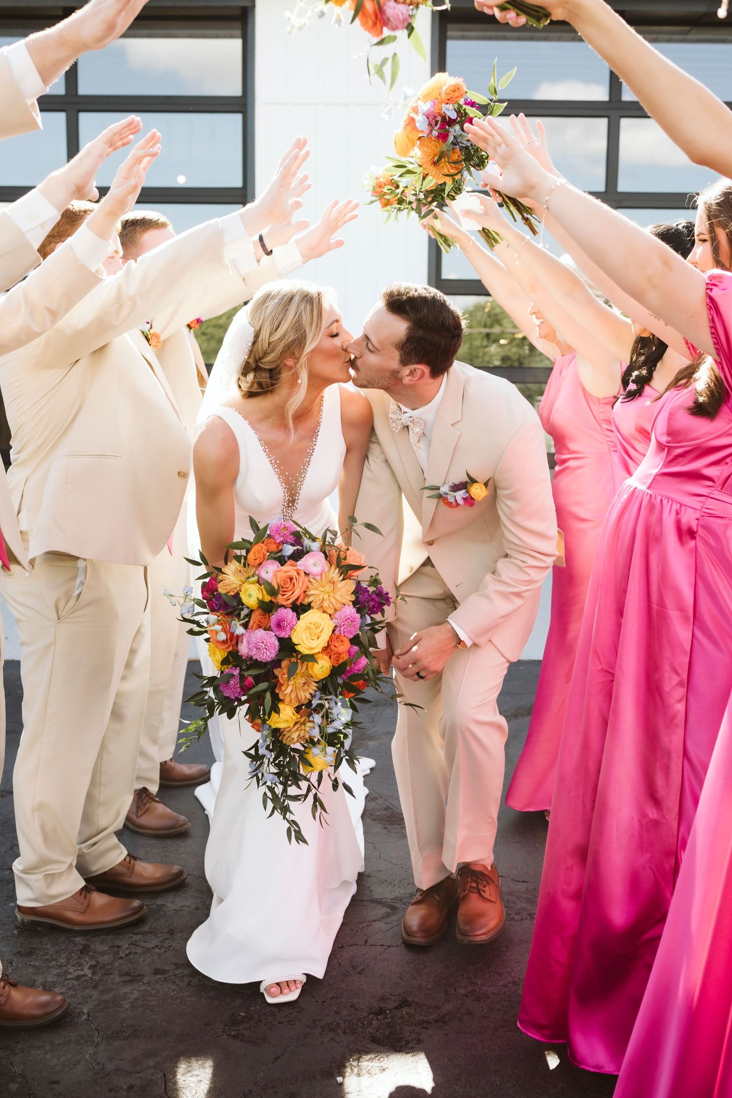 Wedding party forms celebratory arch tunnel as couple shares romantic moment with vibrant bouquet.