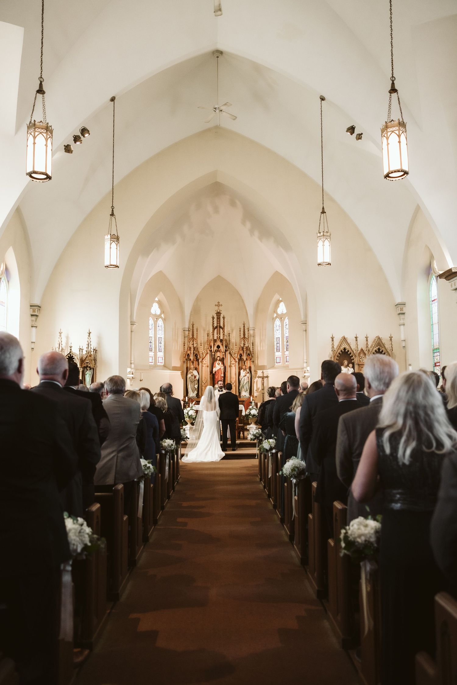 Church filled with wedding guests during ceremony with vaulted ceiling and hanging lights.