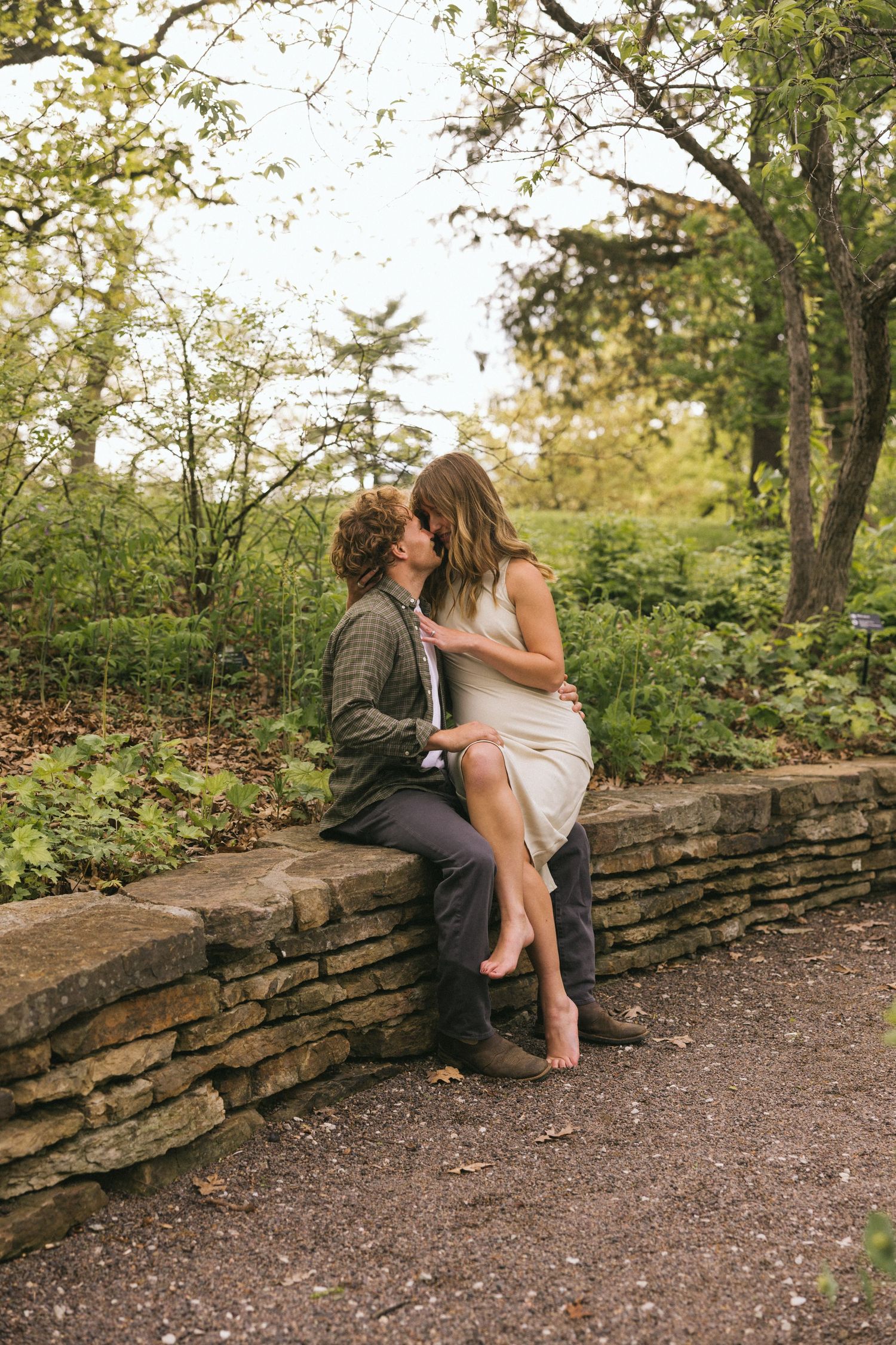 A couple shares intimate moments while sitting on a rustic stone wall surrounded by greenery.