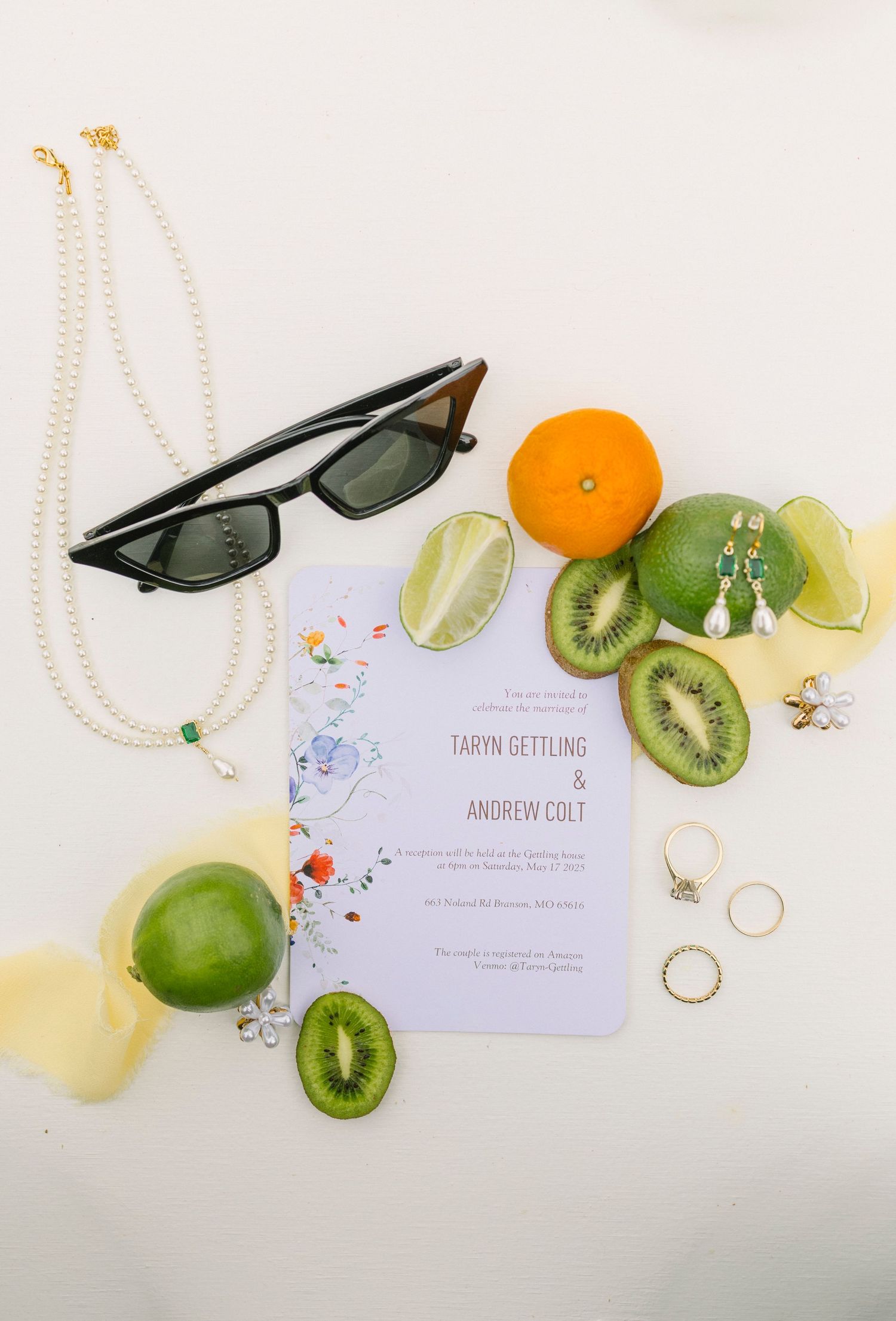 Wedding invitation flatlay with sunglasses, pearl necklace, fresh citrus fruits, kiwi slices and decorative confetti on white.