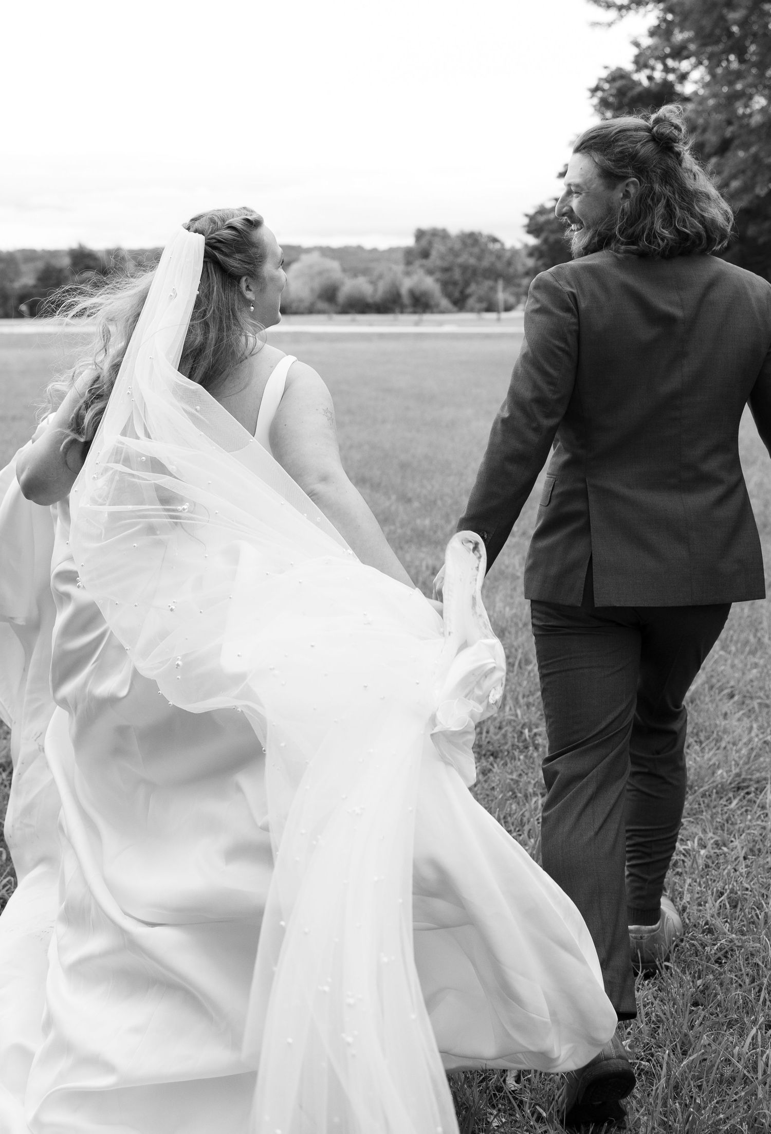 Two figures stroll through a sunlit meadow in black and white with the bride's white flowing dress and veil trailing behind.