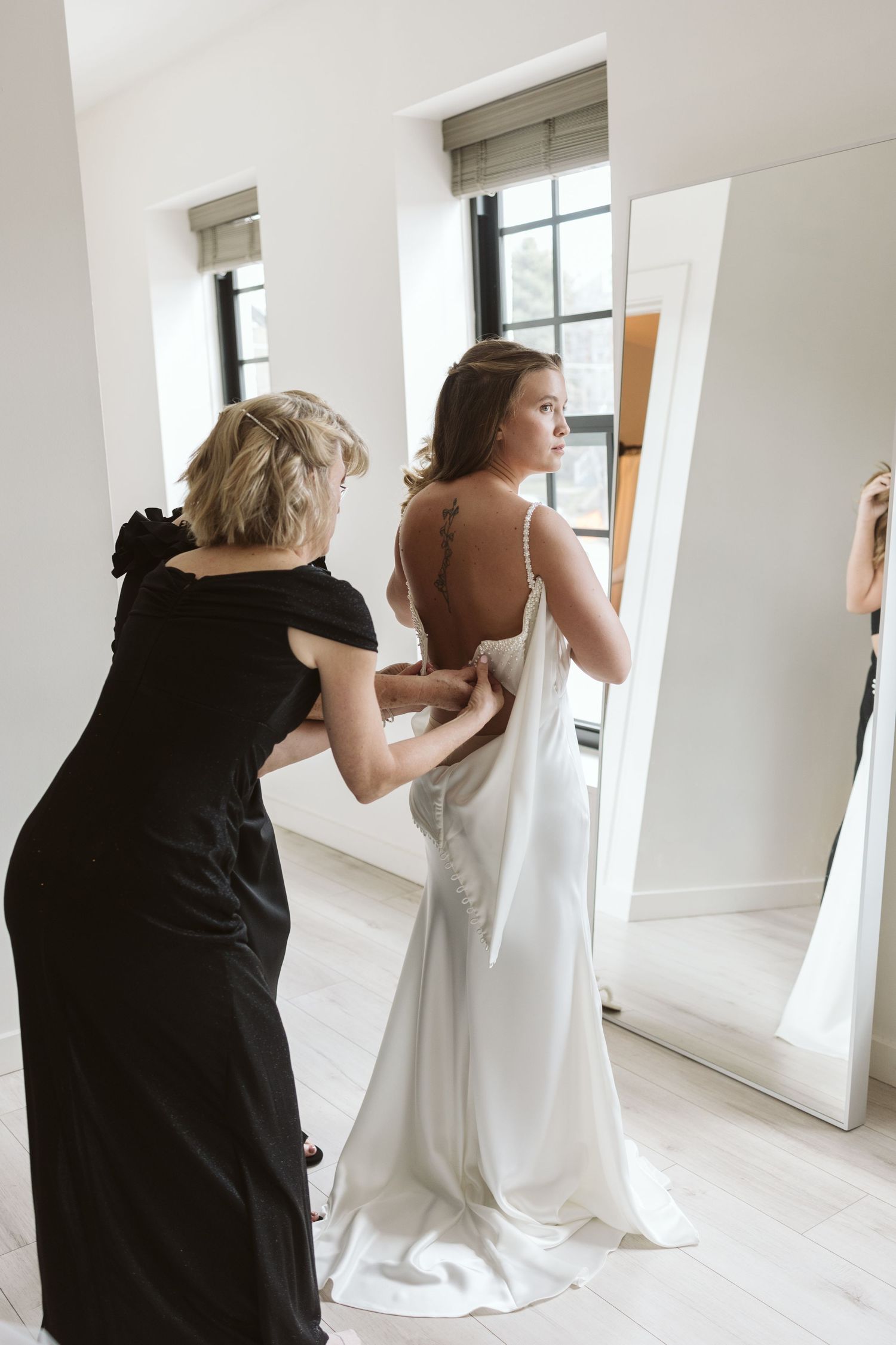 Someone in a black dress helps adjust the back of an elegant white wedding gown in a bright room with large mirrors.