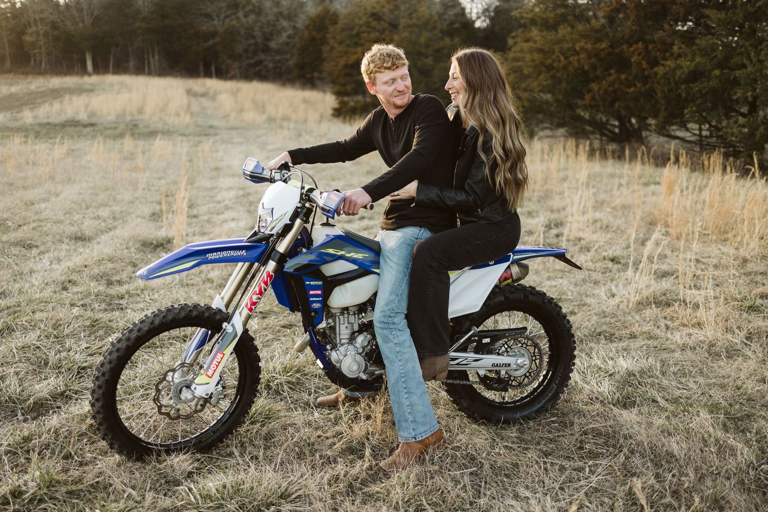 A couple sitting together on a blue dirt bike in a rustic field at sunset.