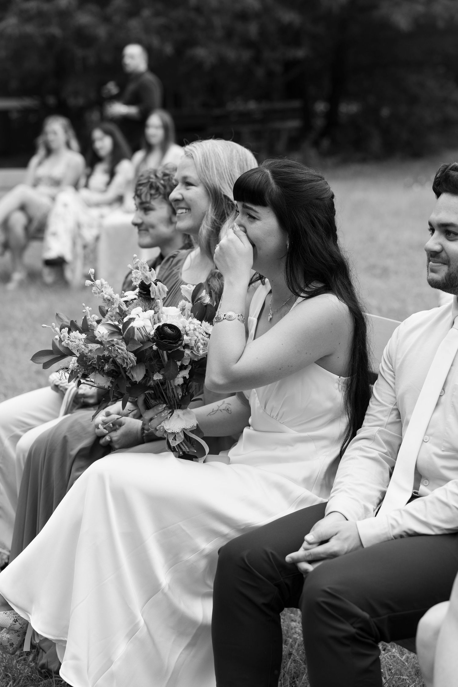Black and white photo of bridesmaids in white dresses seated outdoors holding floral bouquets at a wedding ceremony.