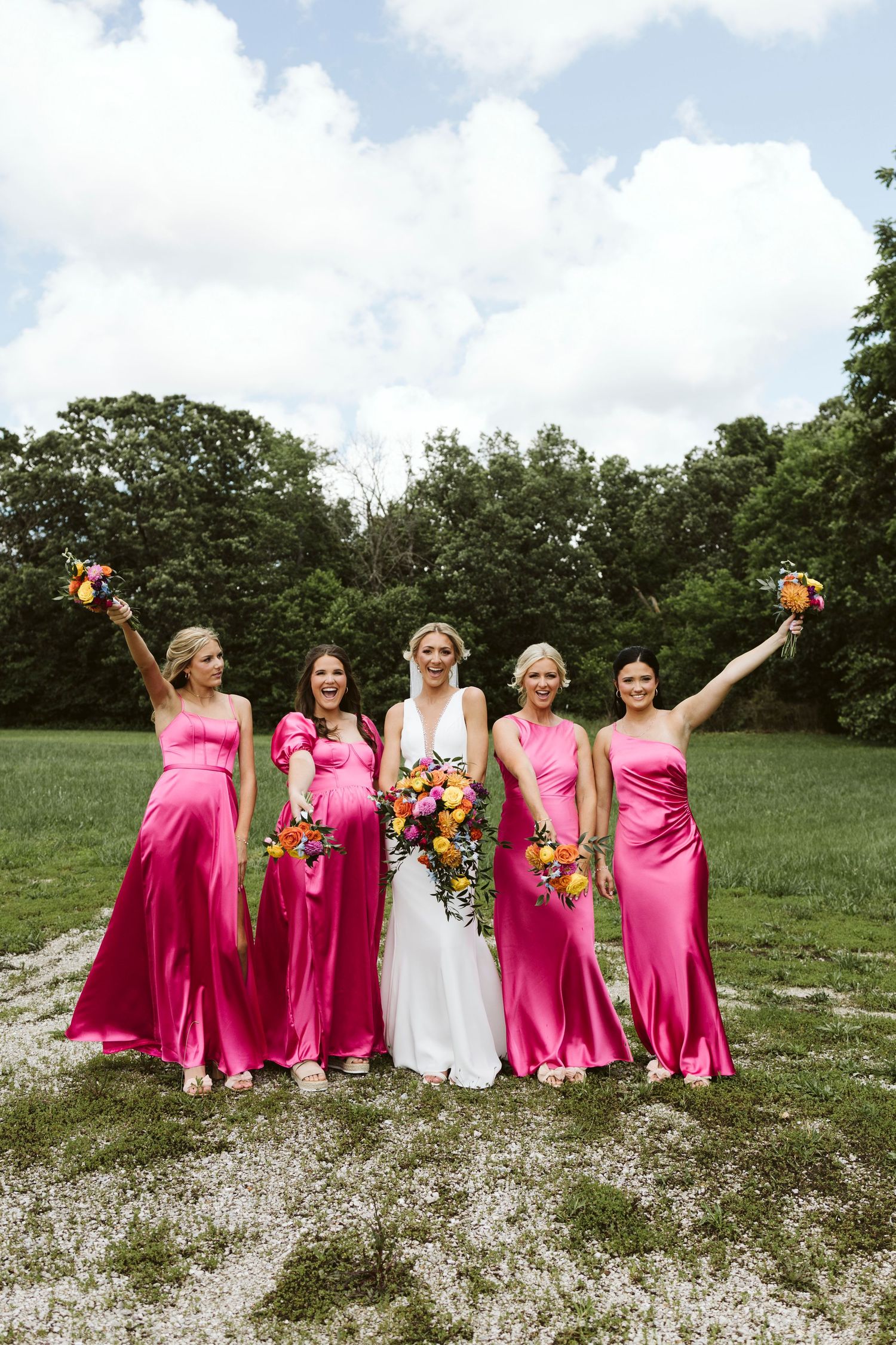 Bridesmaids in bright pink dresses and bride in white pose with bouquets in a sunny outdoor field.