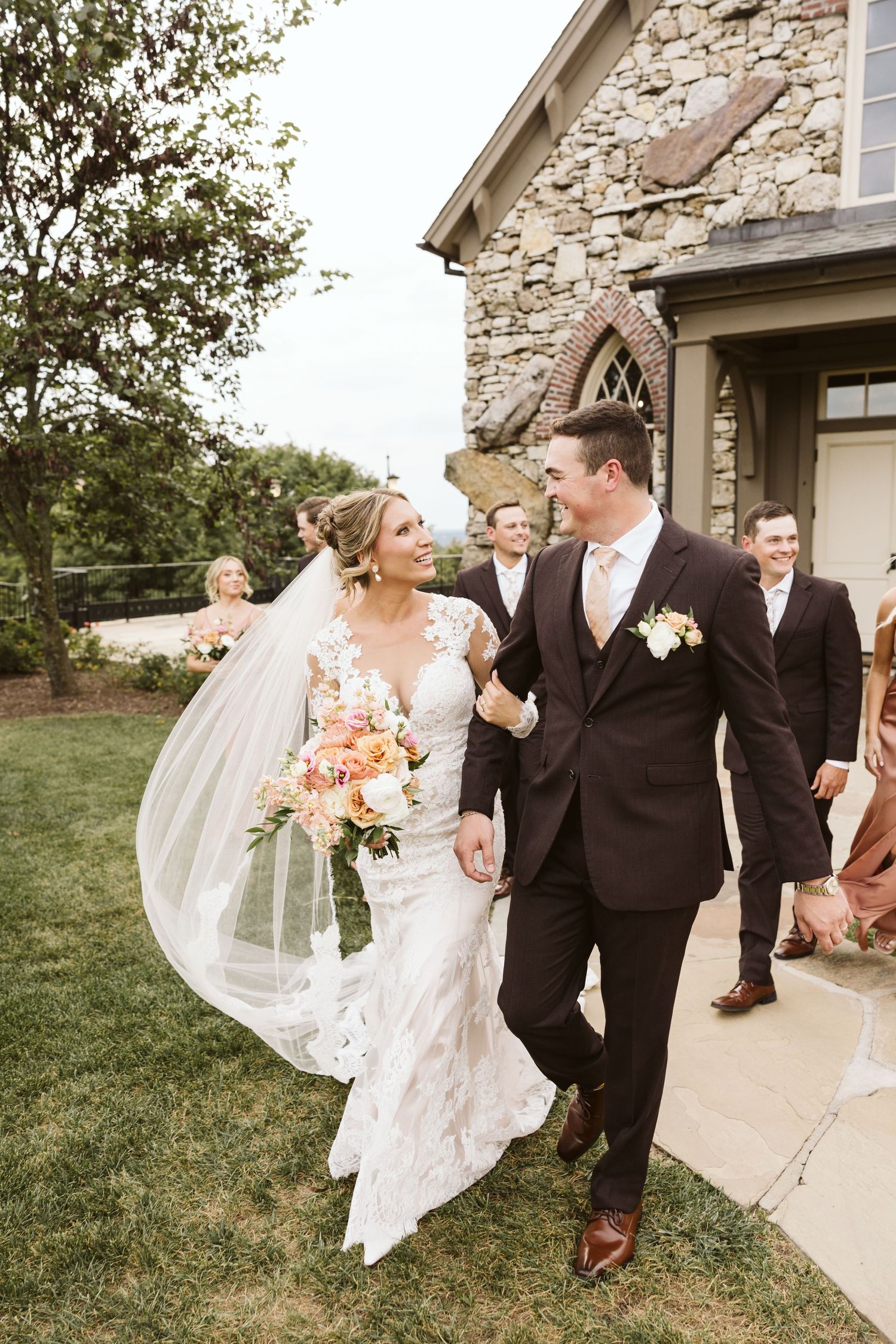 A newly married couple walking outside a stone chapel with wedding guests in the background.