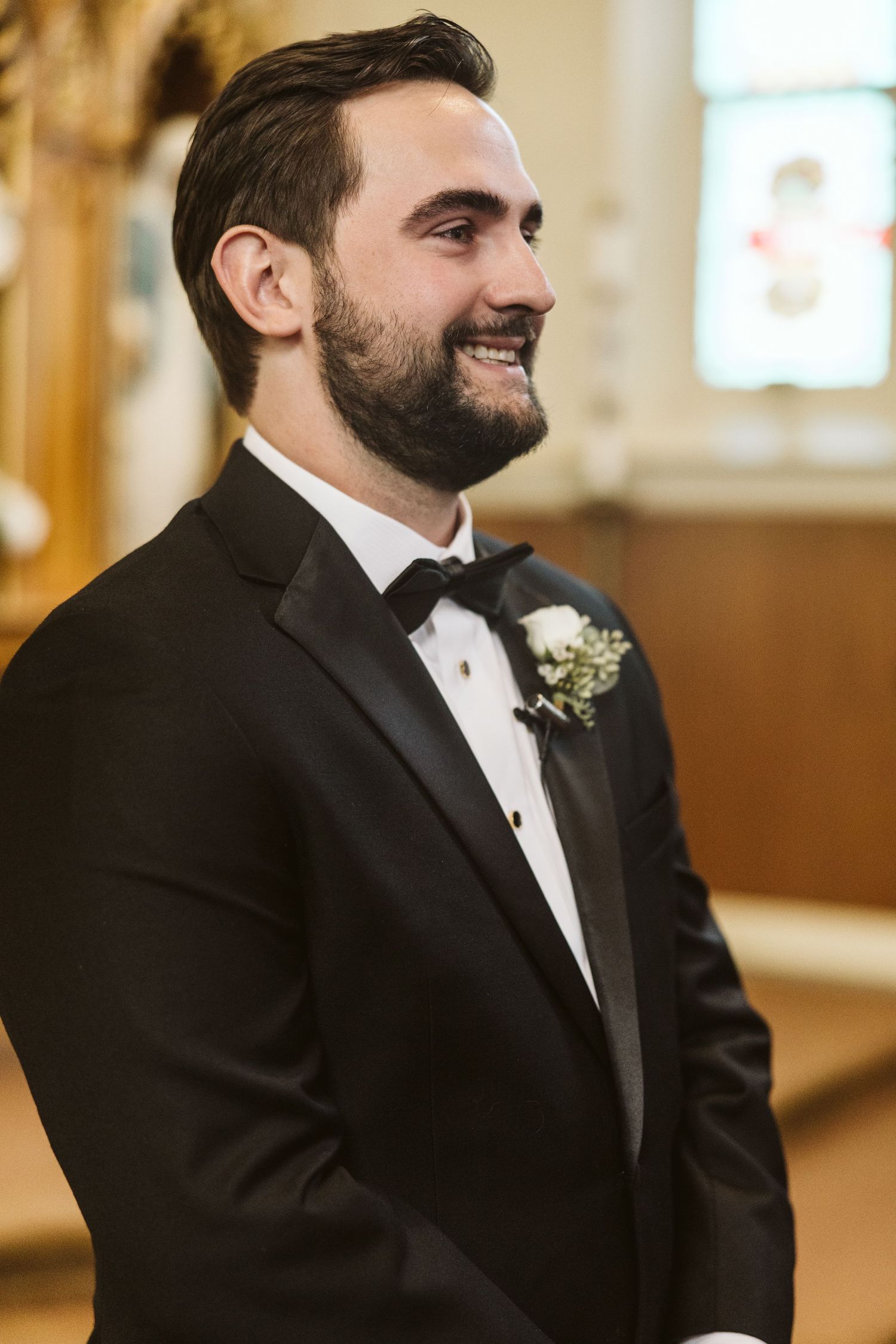 Groom in black tuxedo smiling during wedding ceremony in church.