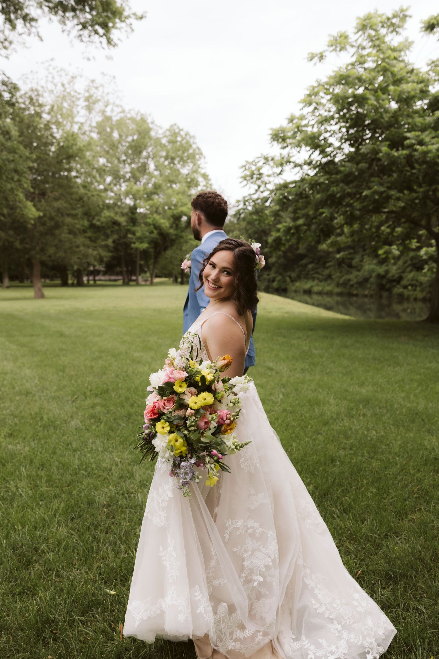 Bride holds colorful bouquet while walking through green meadow with groom in light blue suit.