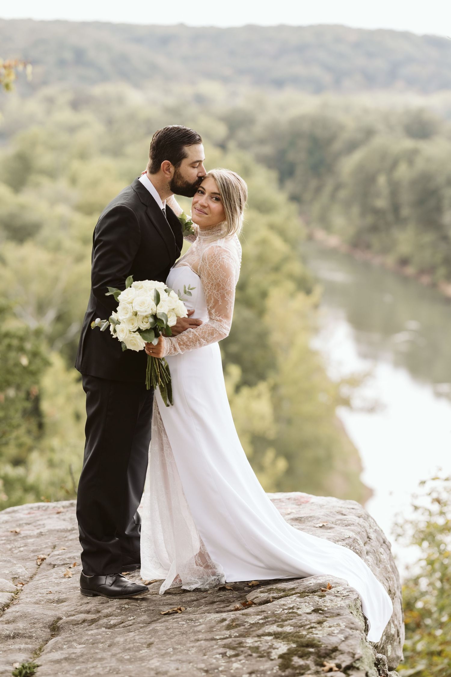 A romantic wedding couple shares a kiss while standing on a rocky overlook above a scenic lake and forest landscape.