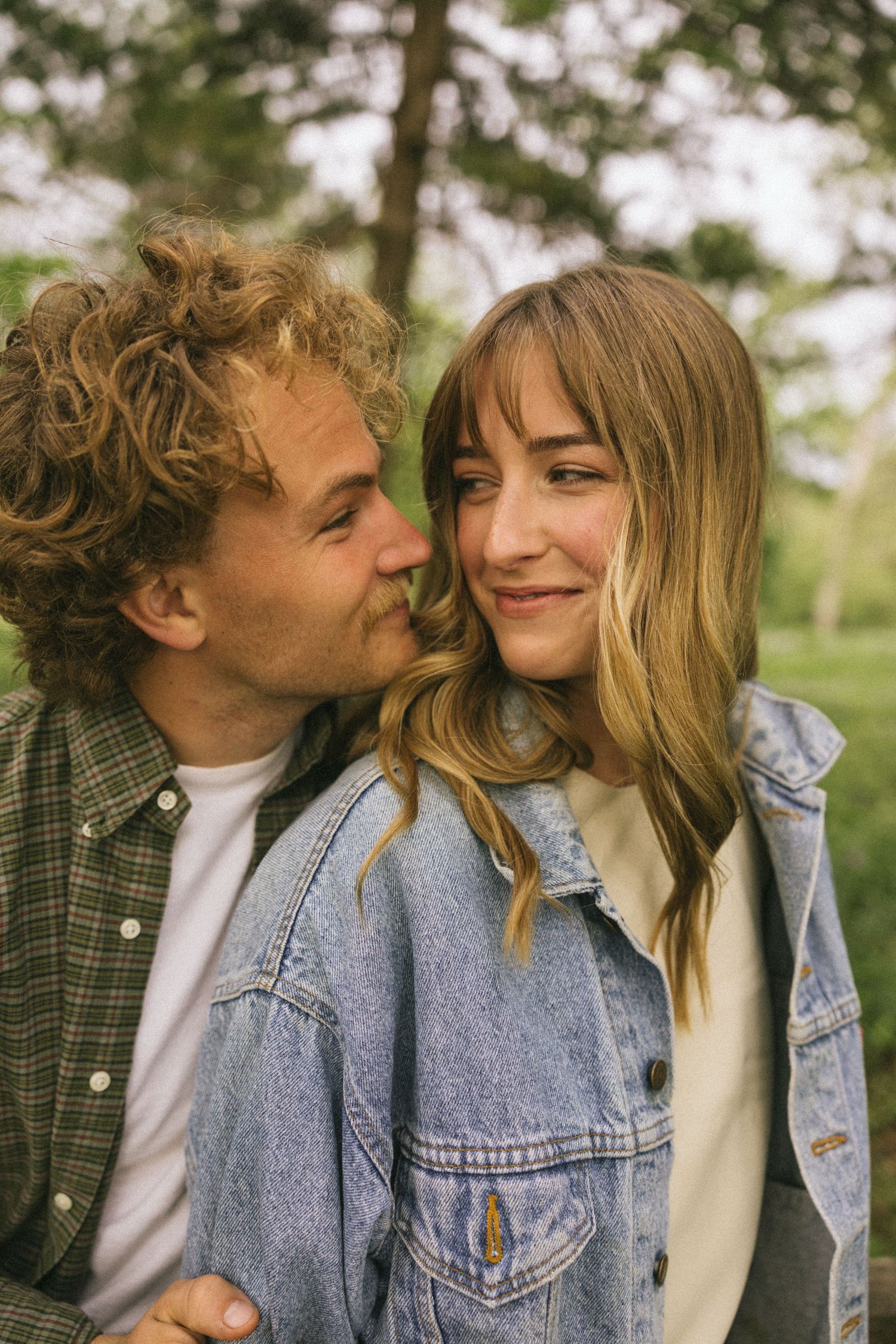 A romantic couple shares intimate moments in a park while wearing casual denim and plaid outfits.