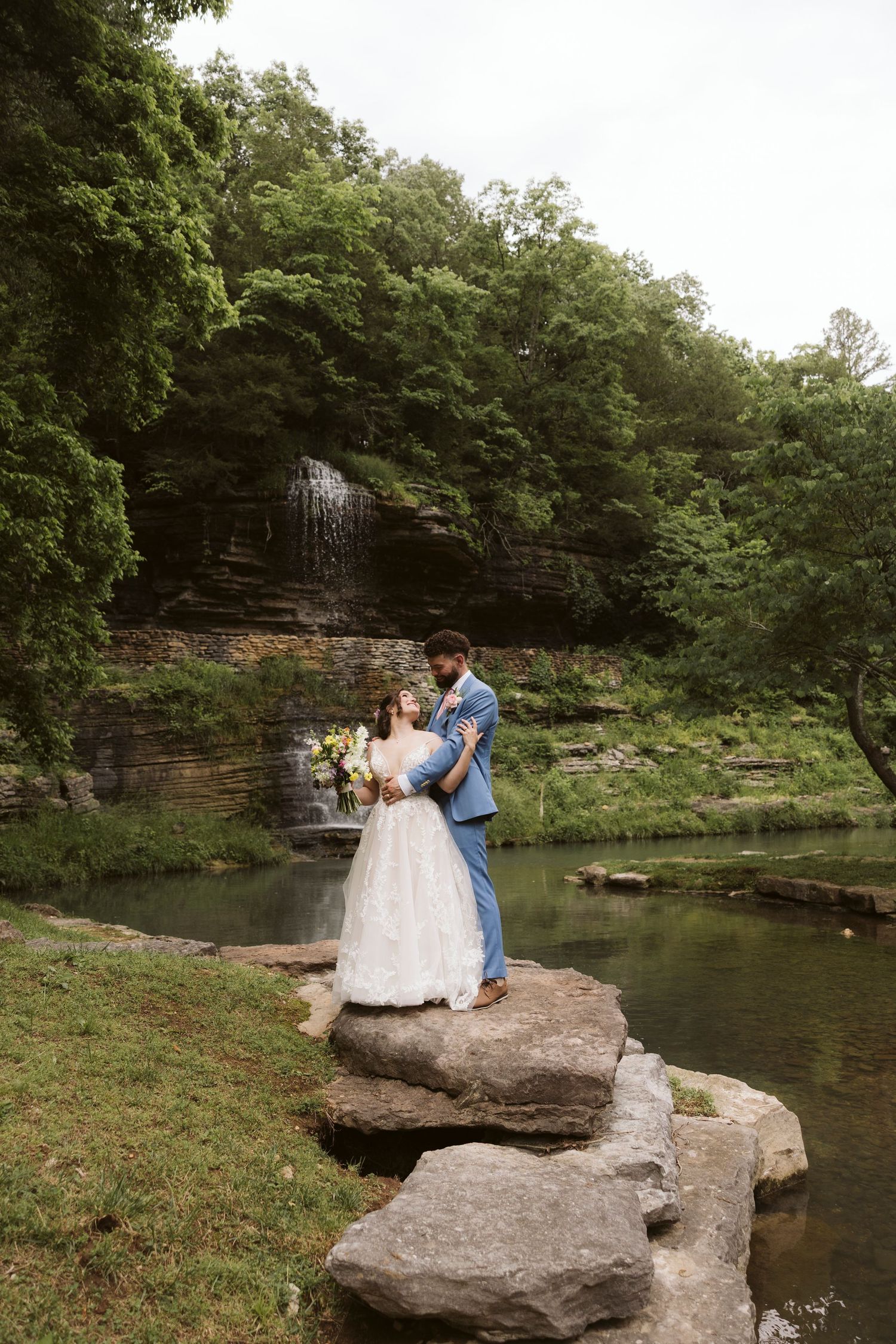 Newlyweds stand on large rocks by a peaceful stream with a cascading waterfall in the background.