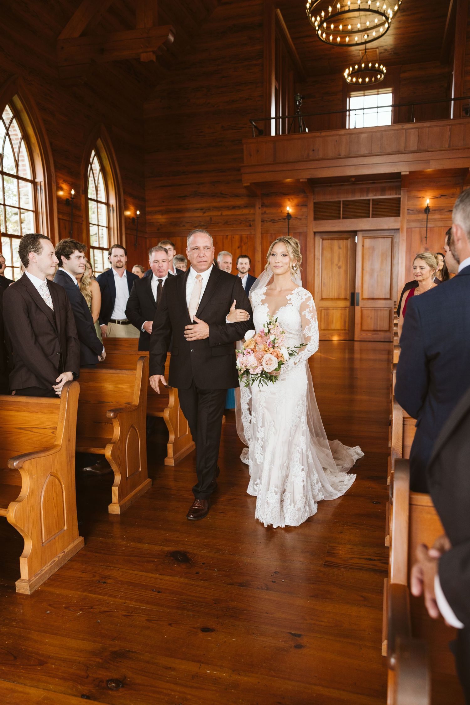 Wedding ceremony processional in wooden chapel with guests seated in pews.
