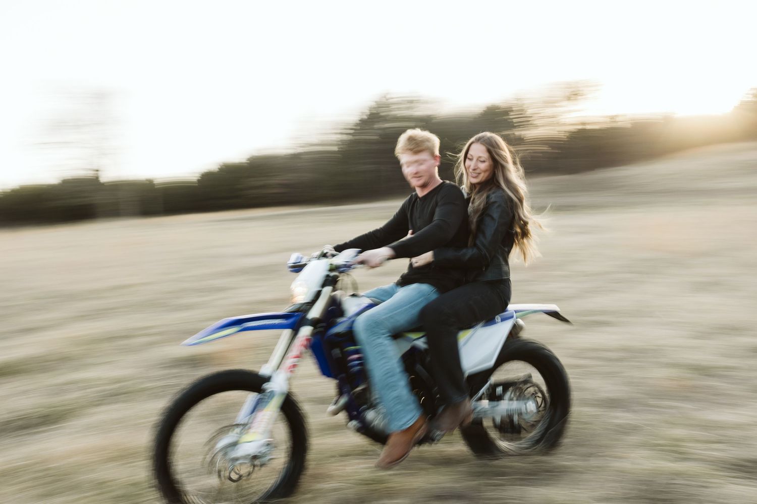 A couple riding together on a blue dirt bike through a field at sunset.