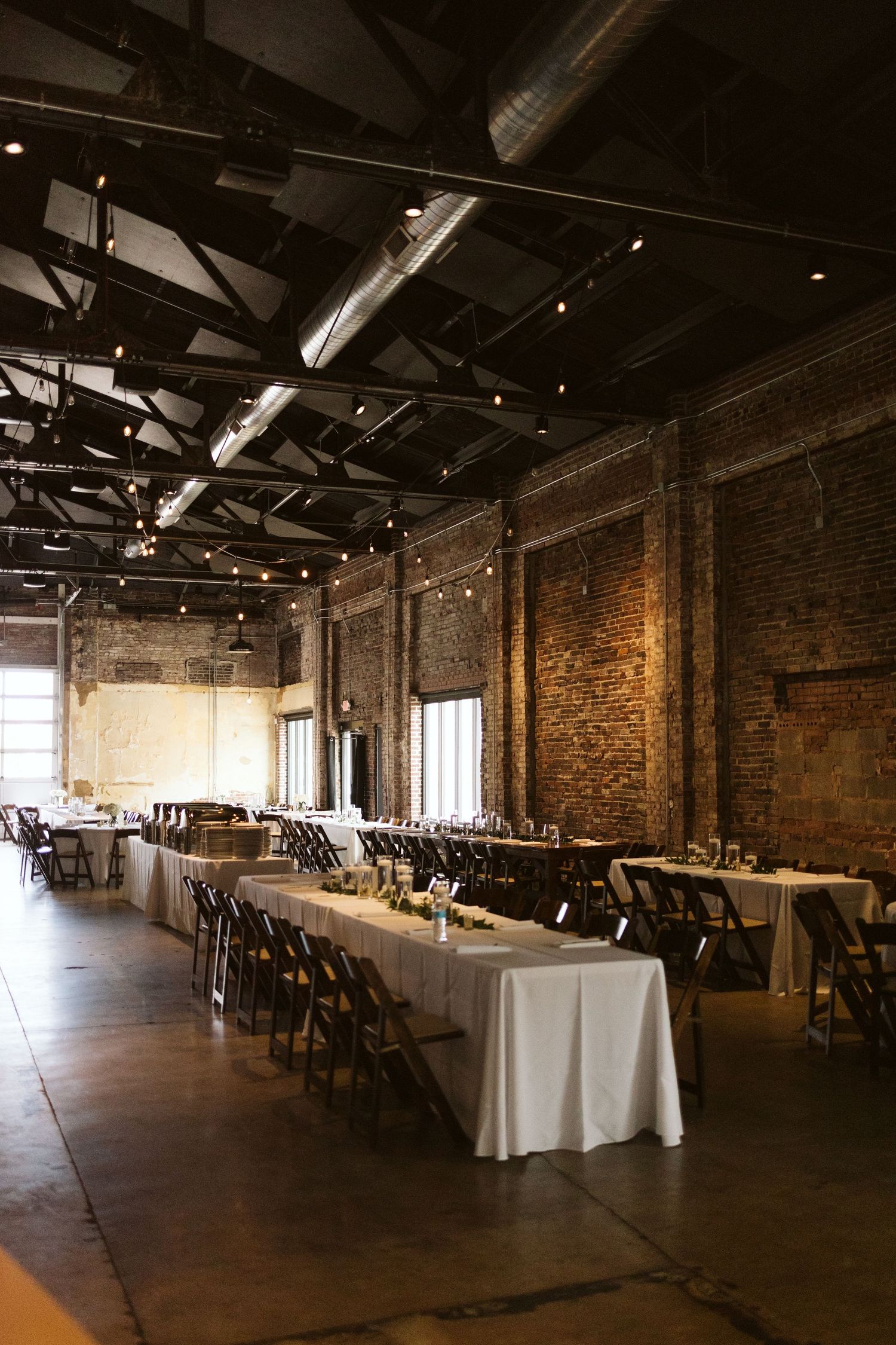 Industrial event space with exposed brick walls, long banquet tables covered in white linens, and string lights overhead.