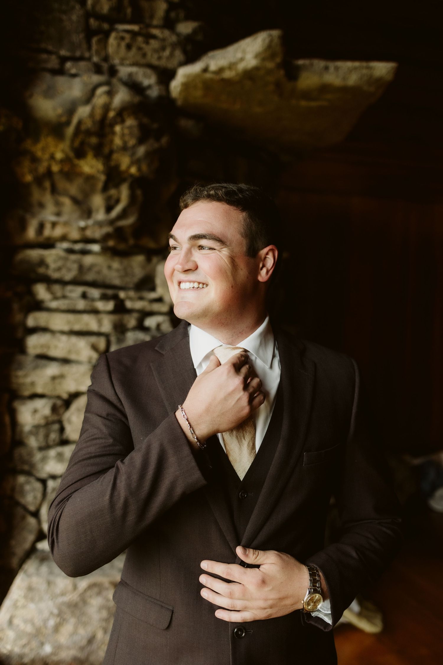 A groom adjusts his tie while smiling in formal attire against a stone wall background.