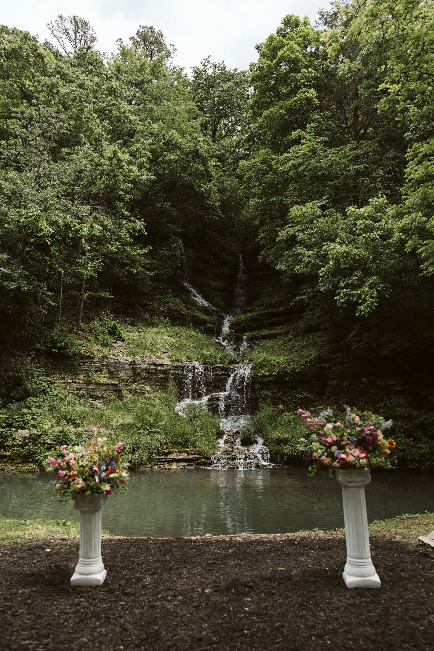 A scenic waterfall backdrop with floral arrangements on pedestals for a wedding ceremony.