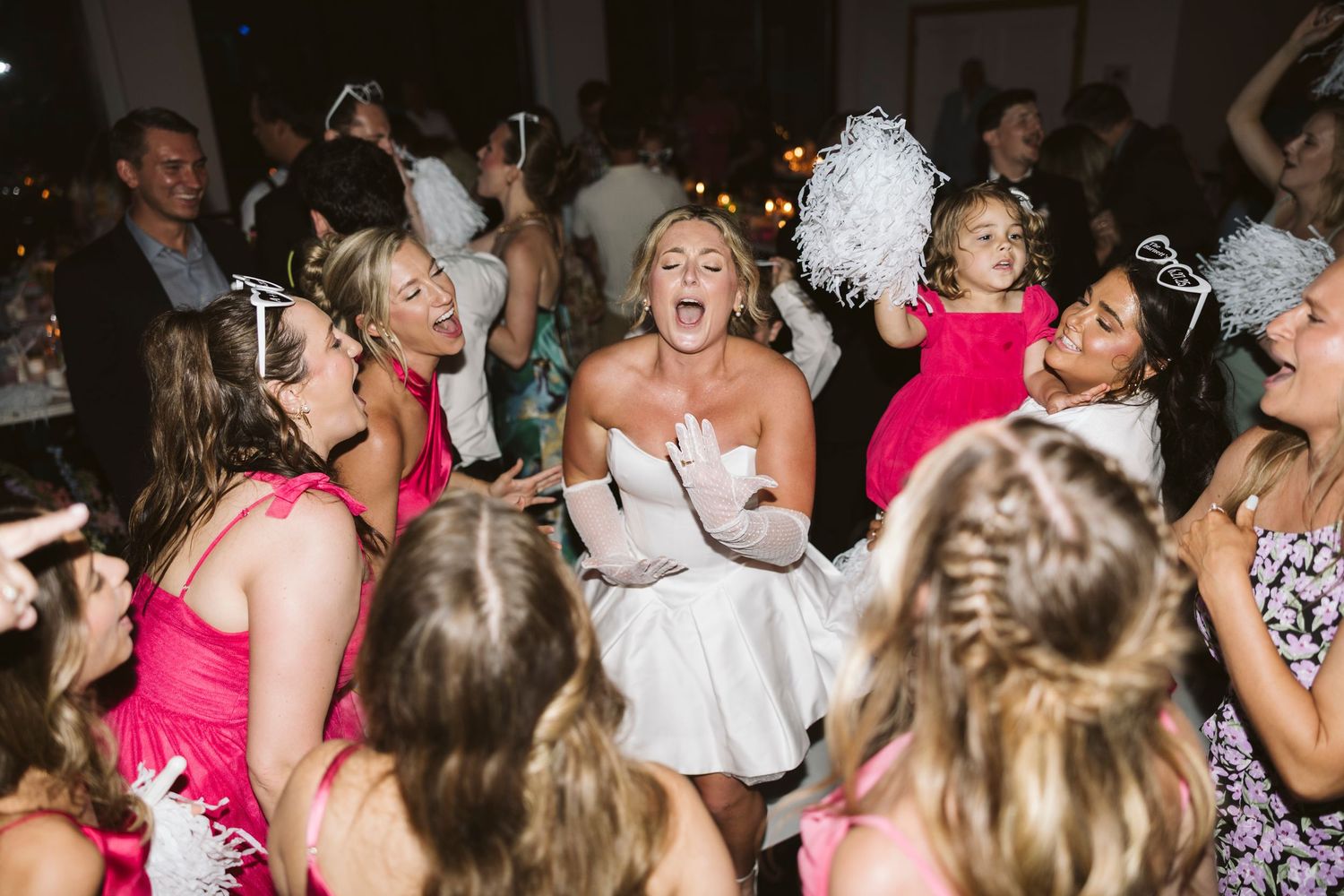 A large crowd of wedding reception guests dance enthusiastically with joyful expressions at a lively party celebration.