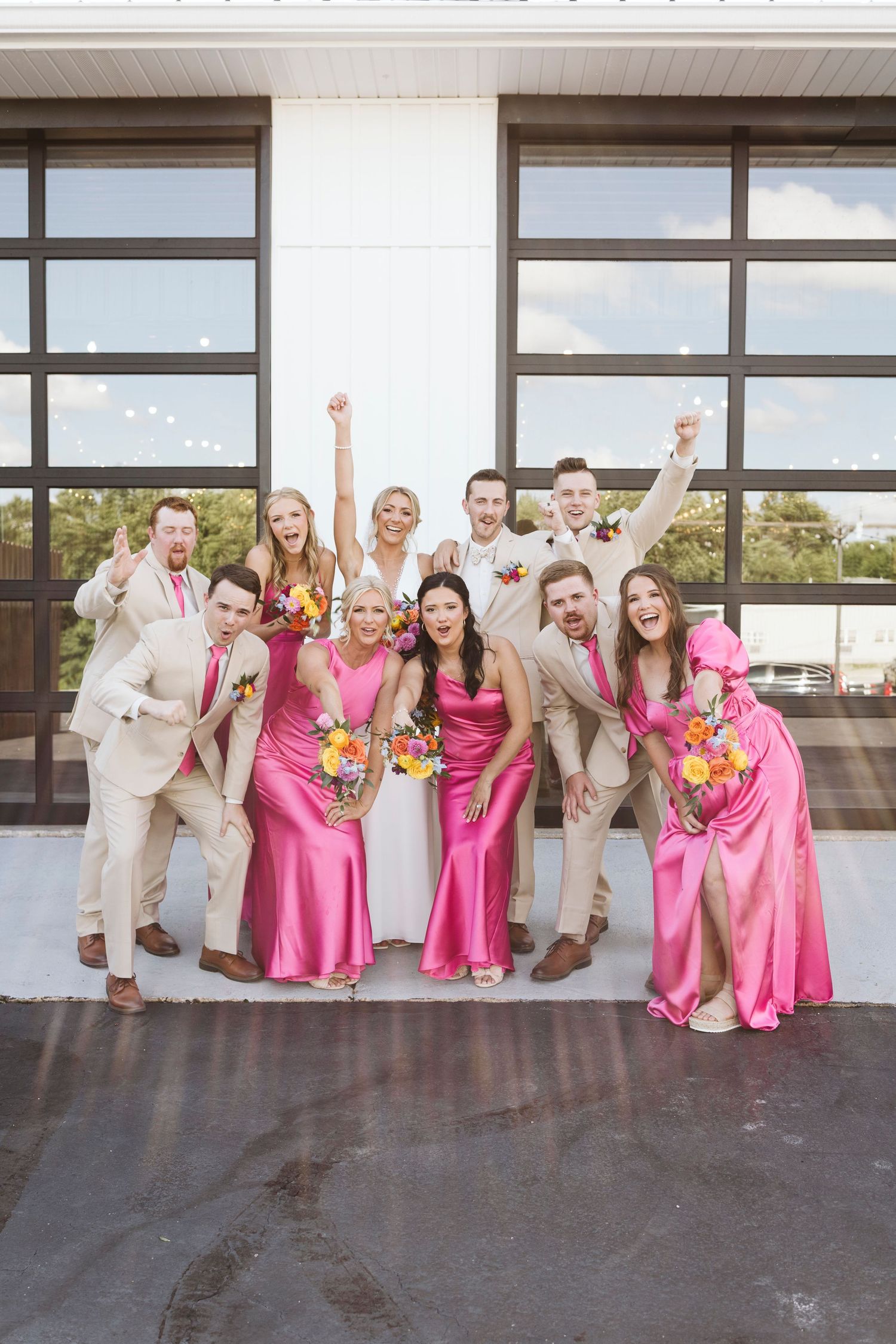 Wedding party in pink bridesmaid dresses and tan suits poses playfully on a rooftop.