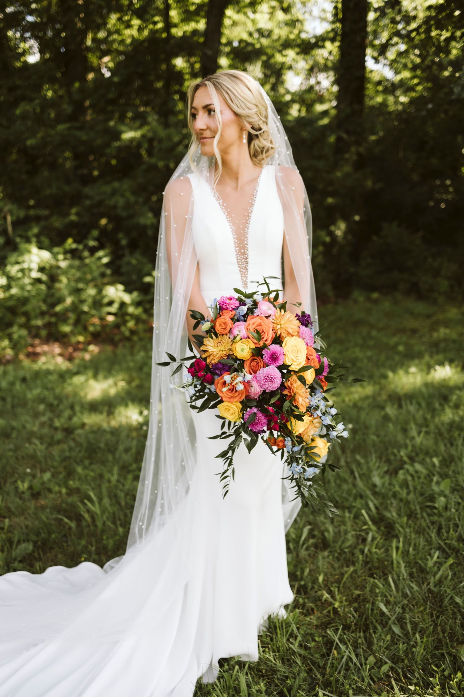 Bride in flowing white gown holds vibrant cascading bouquet of orange and pink flowers in forest setting.
