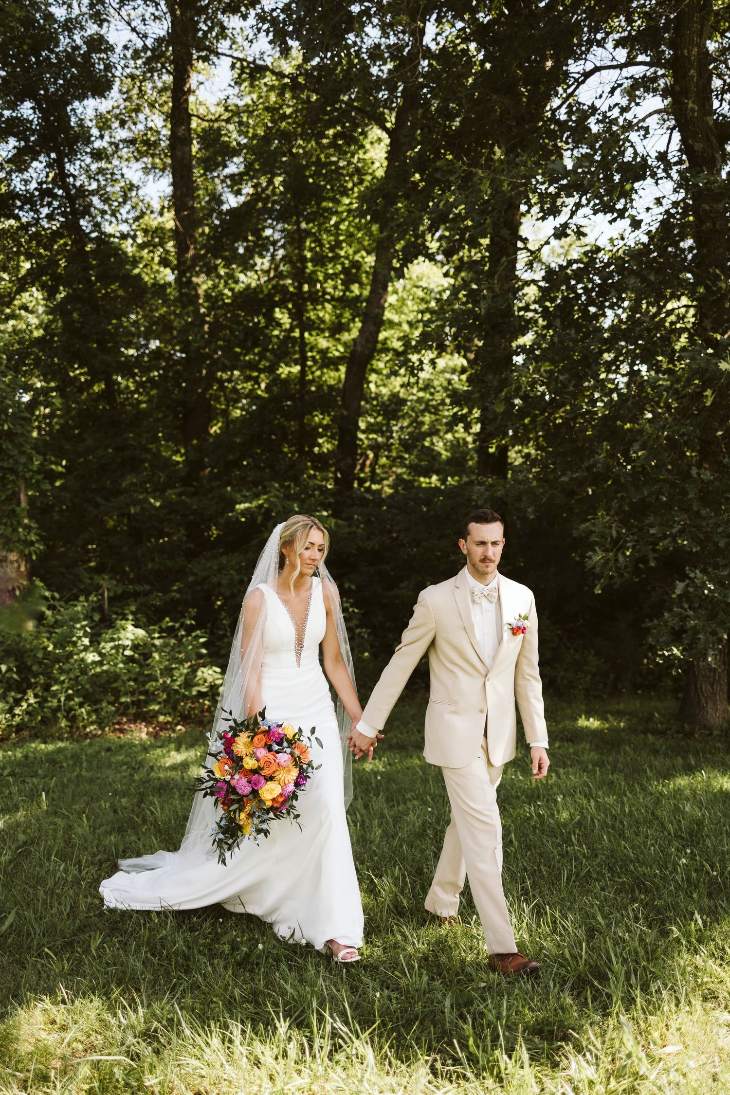 Newlyweds walk hand in hand through sunlit forest, bride carrying vibrant floral bouquet.