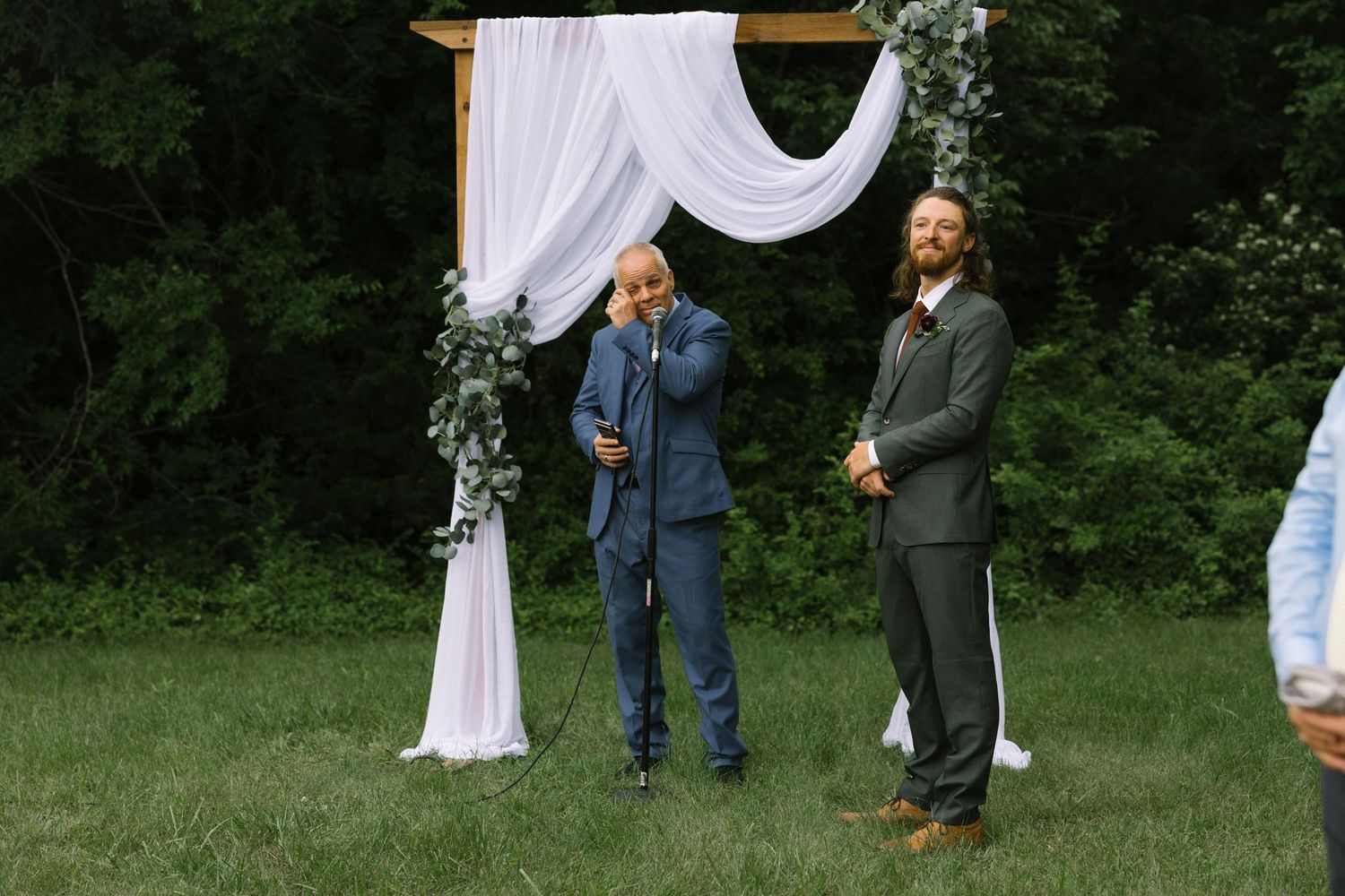An outdoor wedding ceremony setting with a white draped arch and two people standing beneath it in a wooded area.
