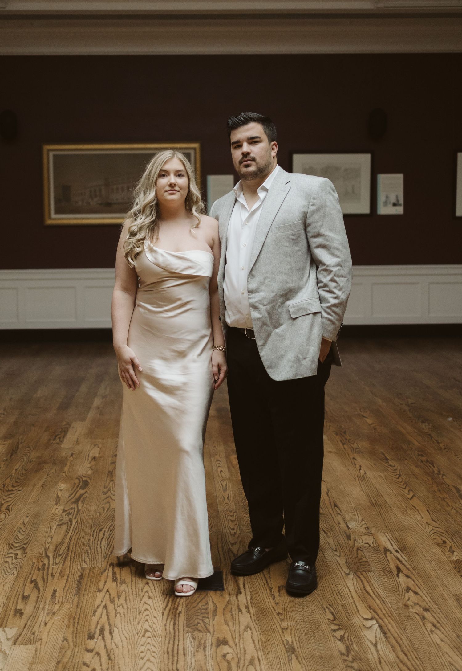 Elegant couple in formal attire standing together in an art gallery with hardwood floors.