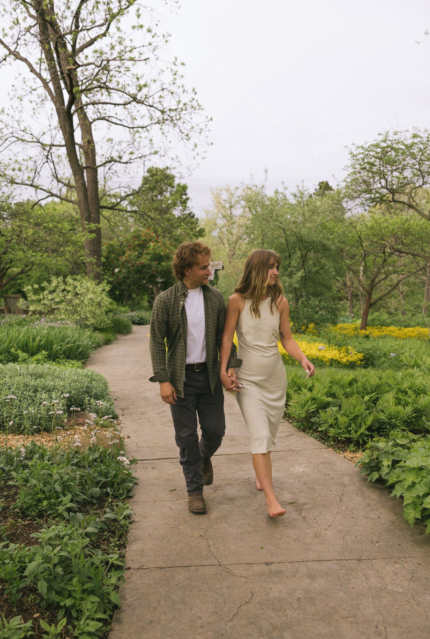 A couple walks hand in hand along a garden path surrounded by lush greenery and yellow flowers.