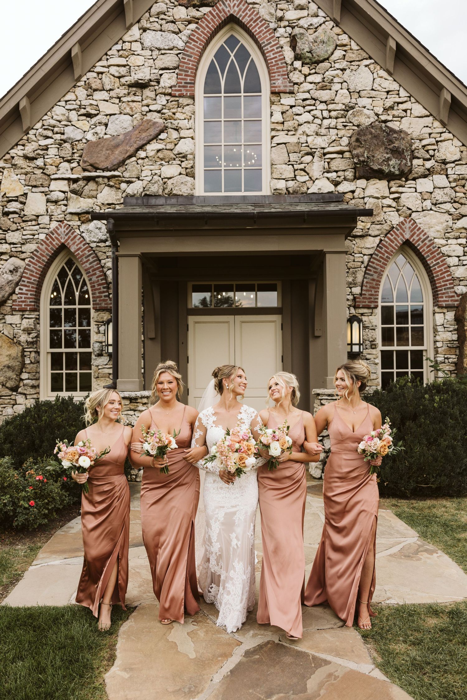 Bridal party in dusty rose dresses posing in front of a stone chapel with Gothic windows.