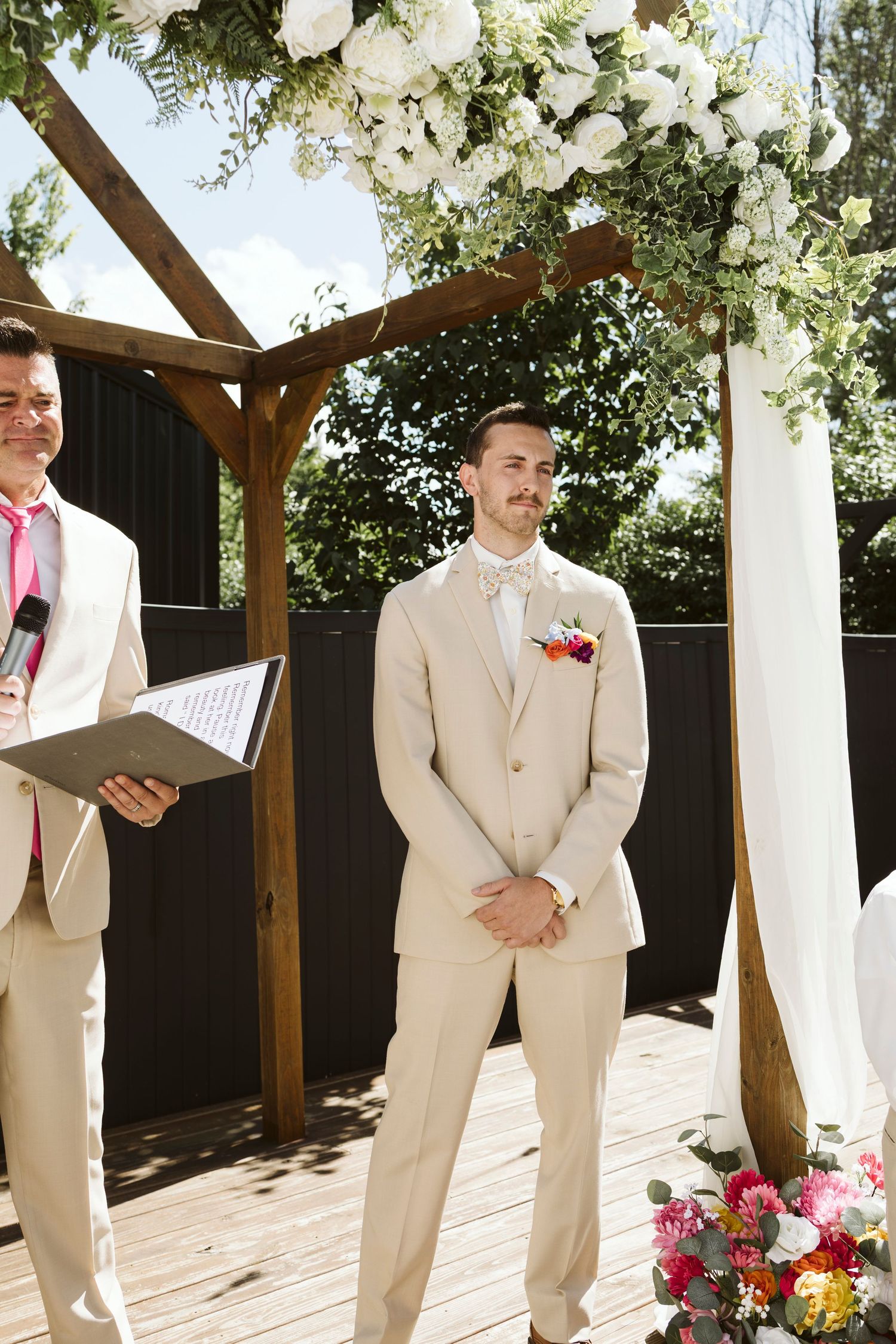 Groomsmen in tan suits stand under a wooden arch decorated with white floral arrangements.