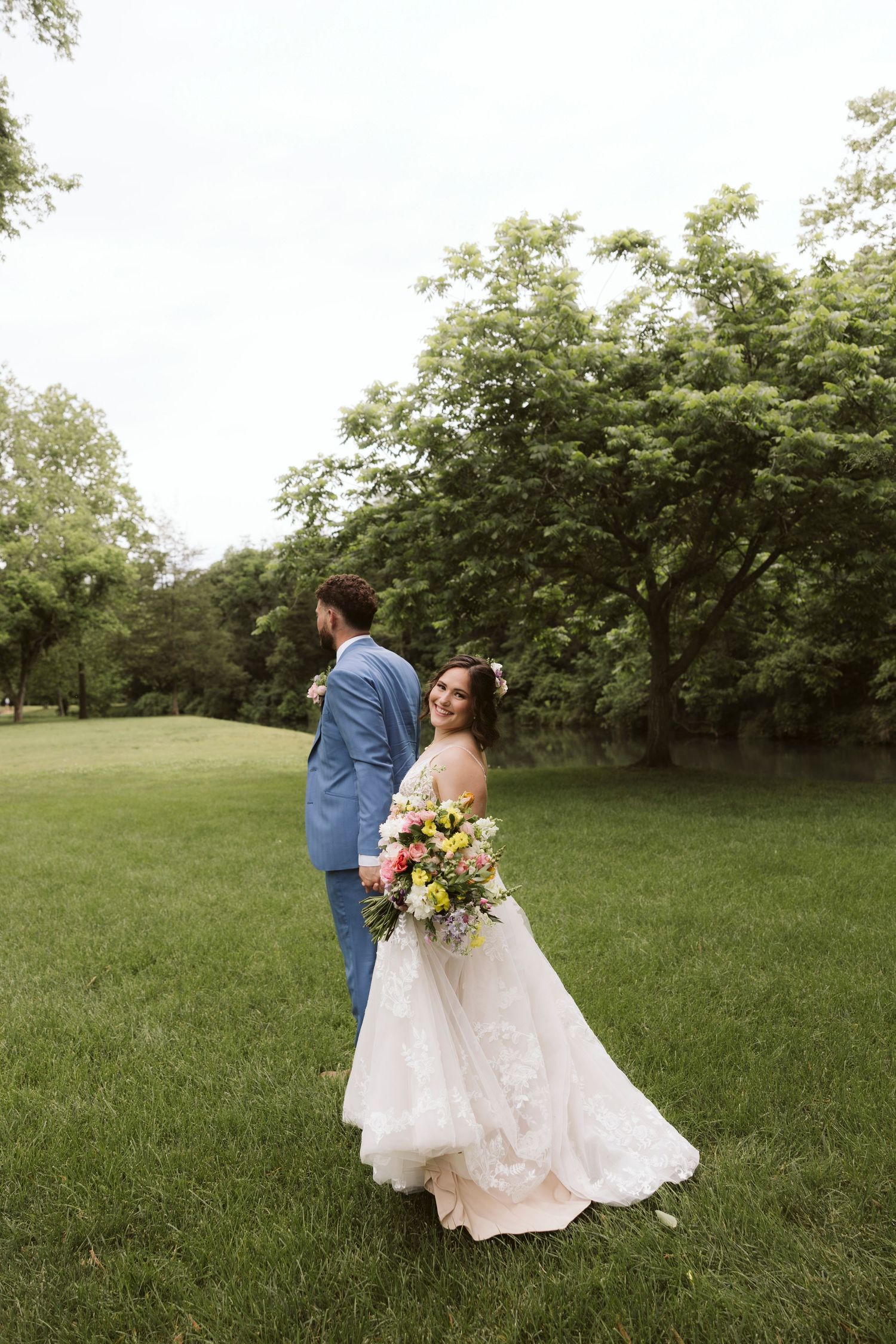 A couple in wedding attire walks through a lush green garden during their wedding photoshoot.