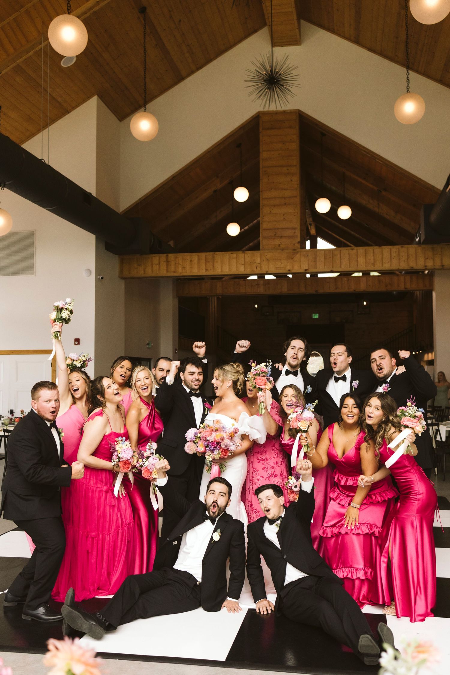 A wedding party in bright pink bridesmaids dresses and black tuxedos poses together in a venue with wooden beams.
